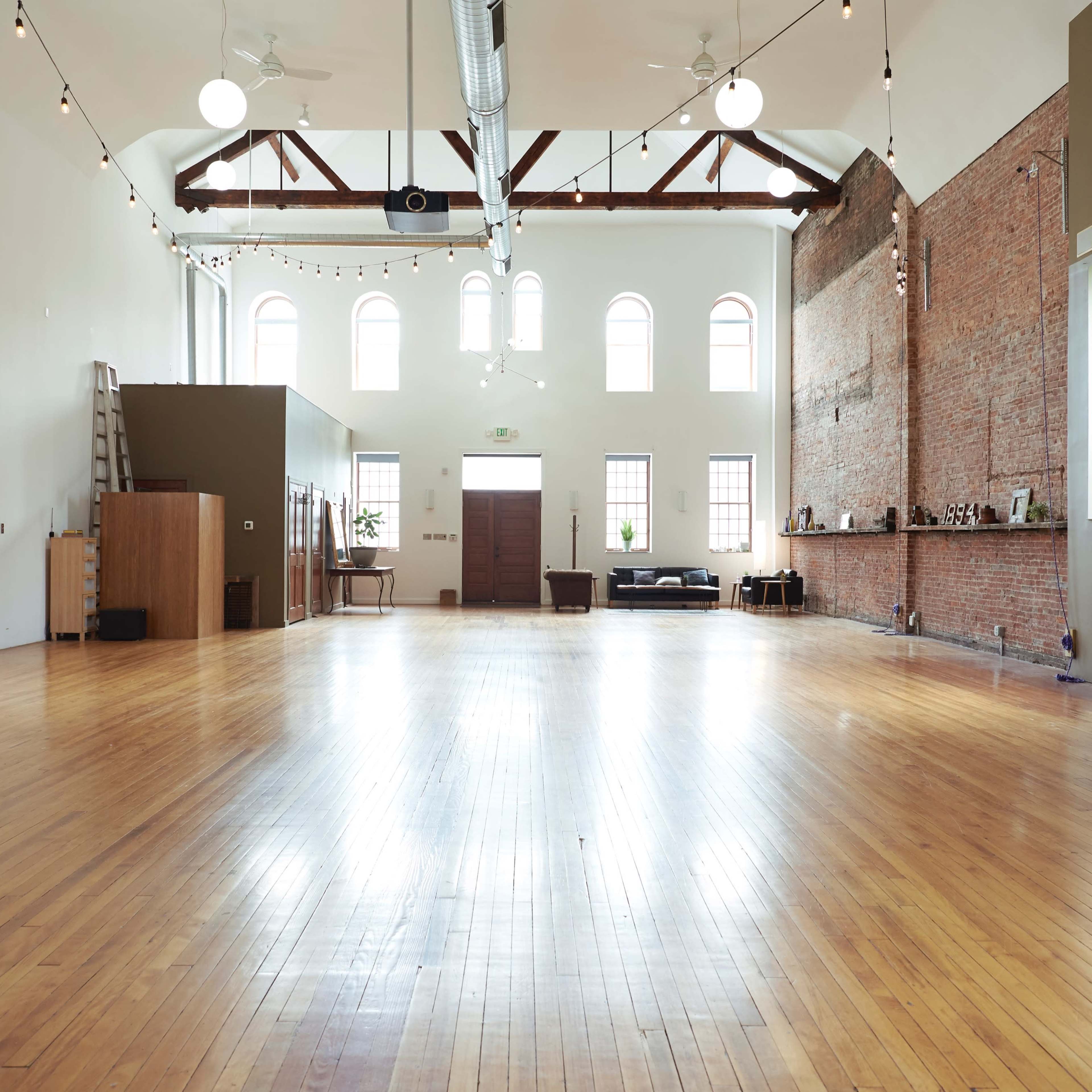 A spacious, wooden-floored room with high ceilings, large windows, and a mix of brick and smooth walls.