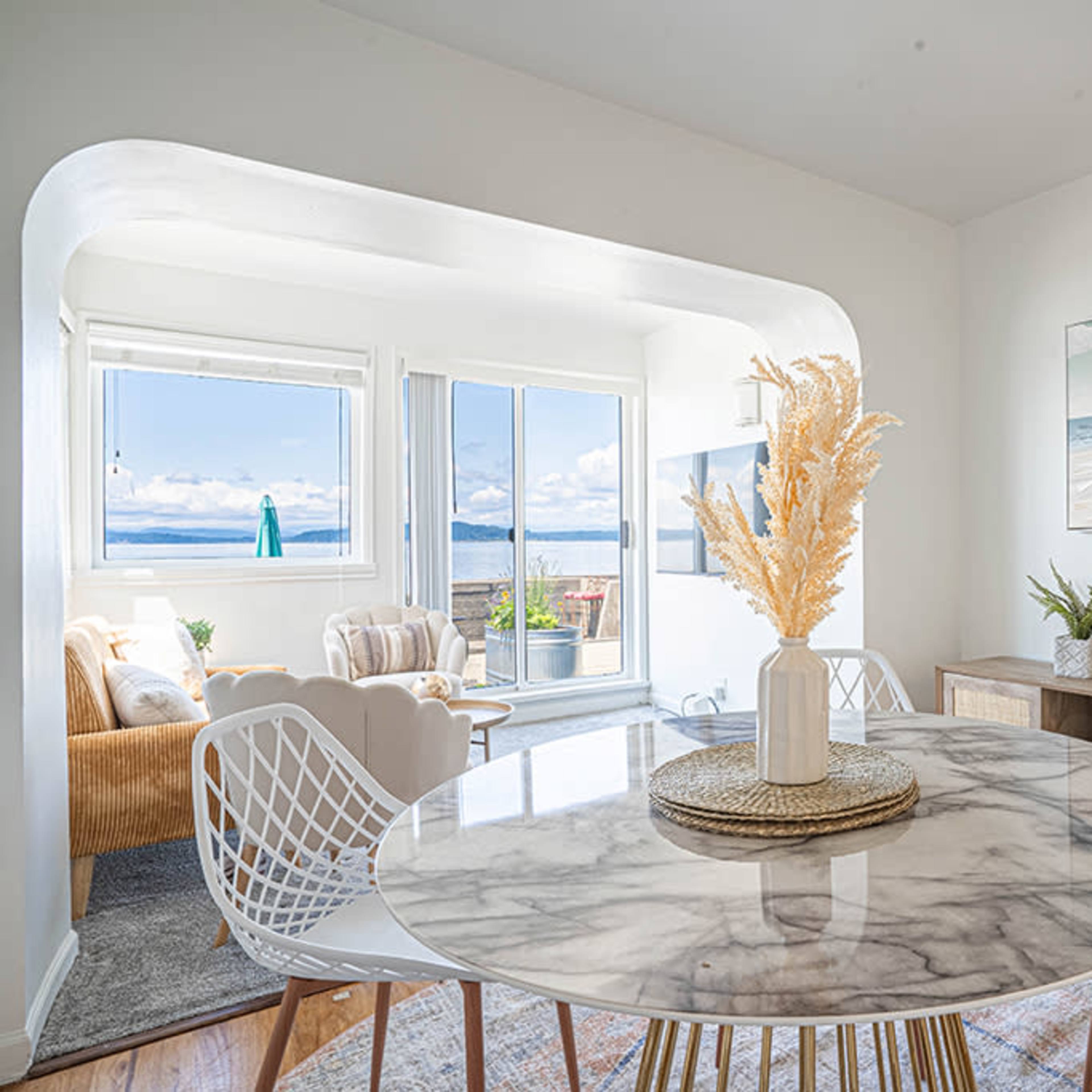 A modern dining area features a round marble table with white chairs, complemented by a decorative vase and a view of the ocean through large windows.