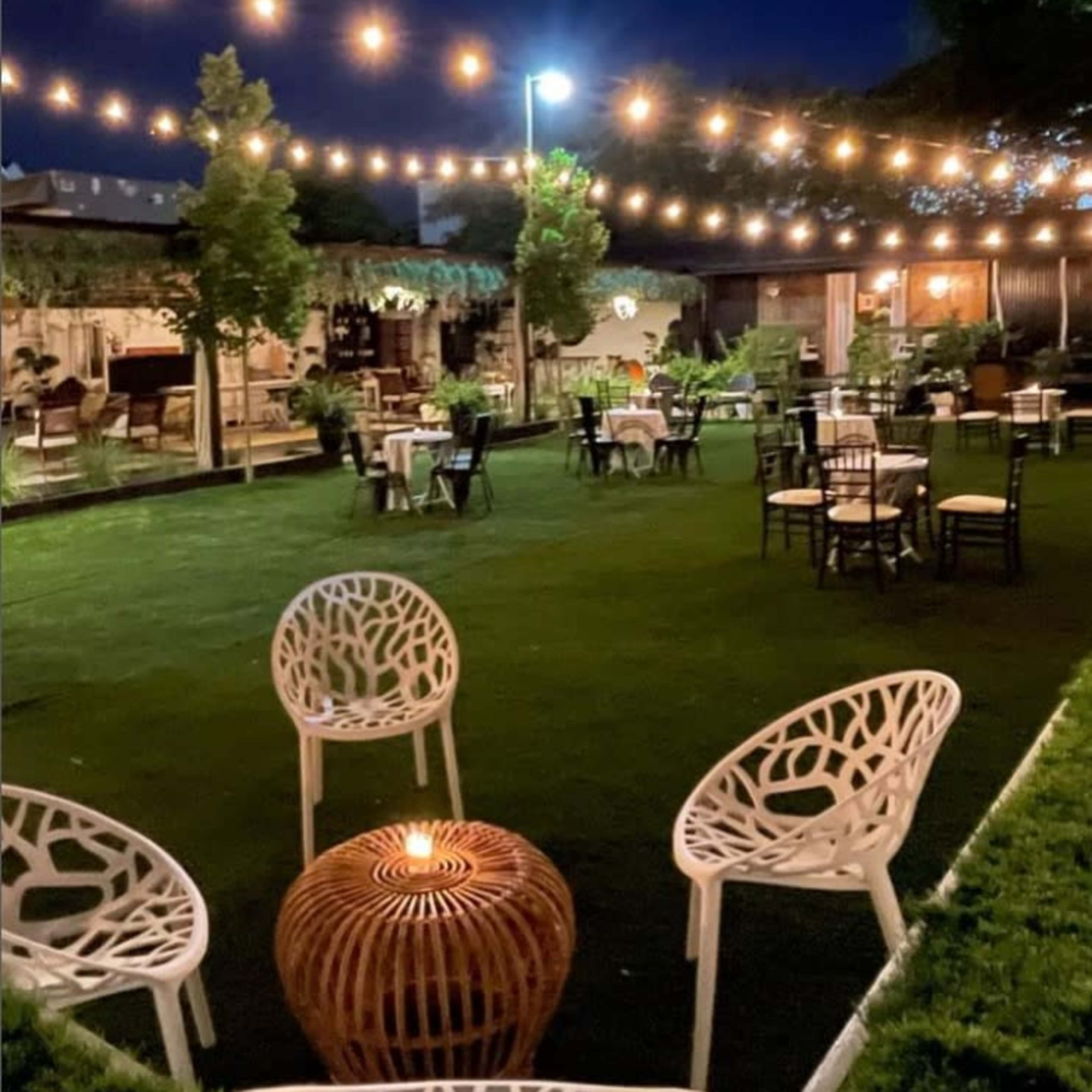 An outdoor restaurant with string lights overhead, featuring green grass, circular tables, and white chairs arranged around a central fire pit.