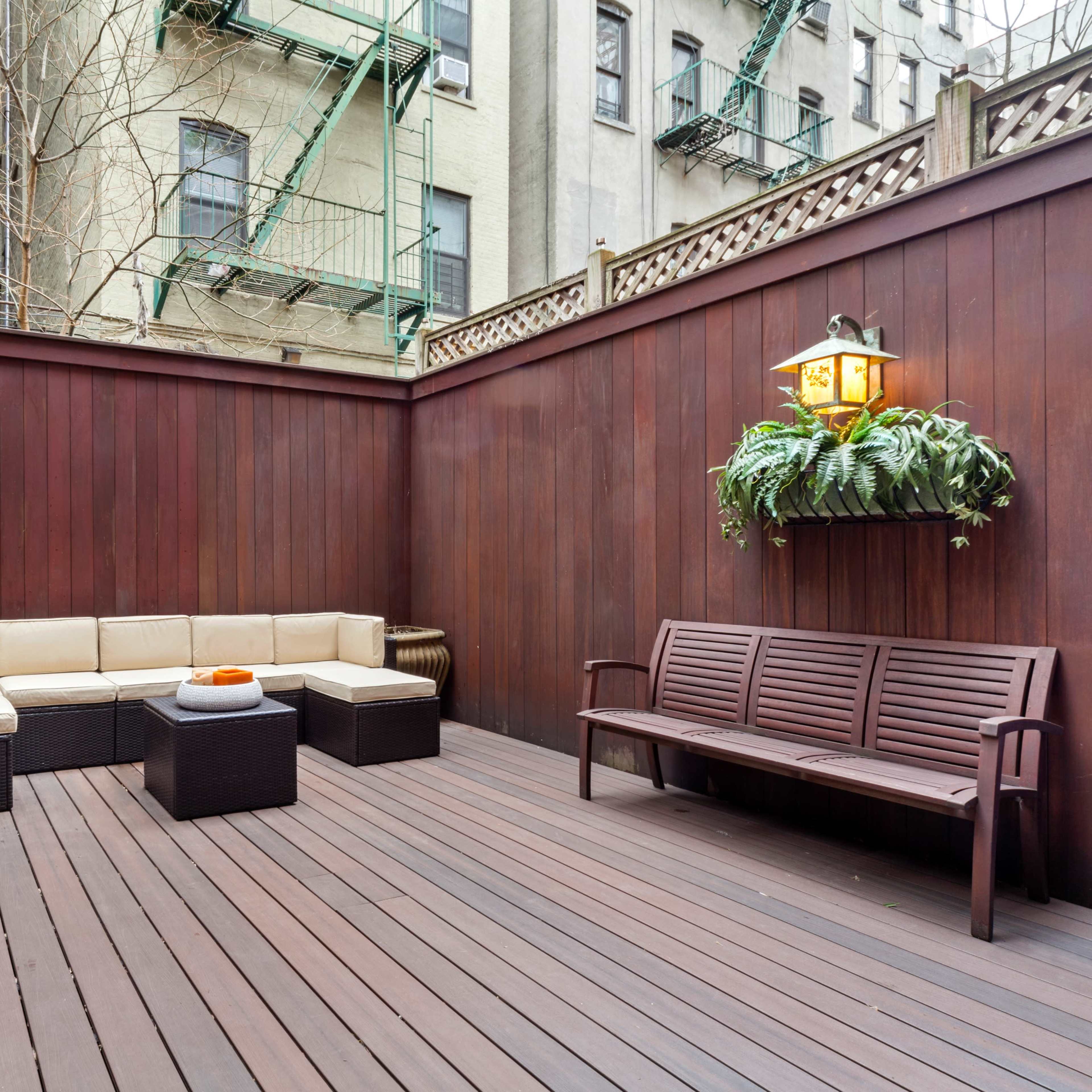 A small outdoor patio surrounded by wooden walls, featuring a seating area with a couch, a coffee table, and a bench, along with hanging plants.
