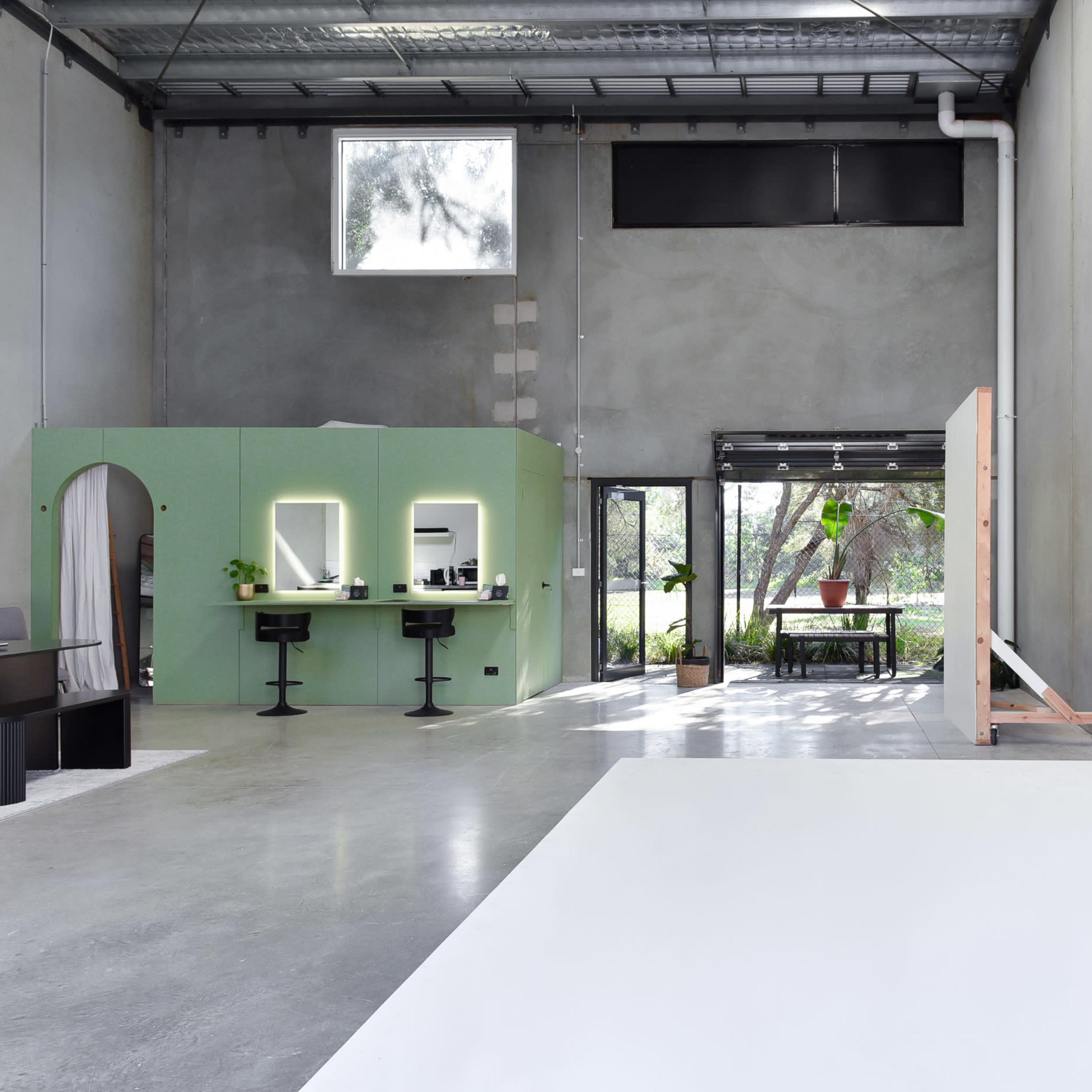 A spacious, industrial-style interior with a green partition housing mirrors and black stools, leading to an area with a large white backdrop and natural light coming through large windows.