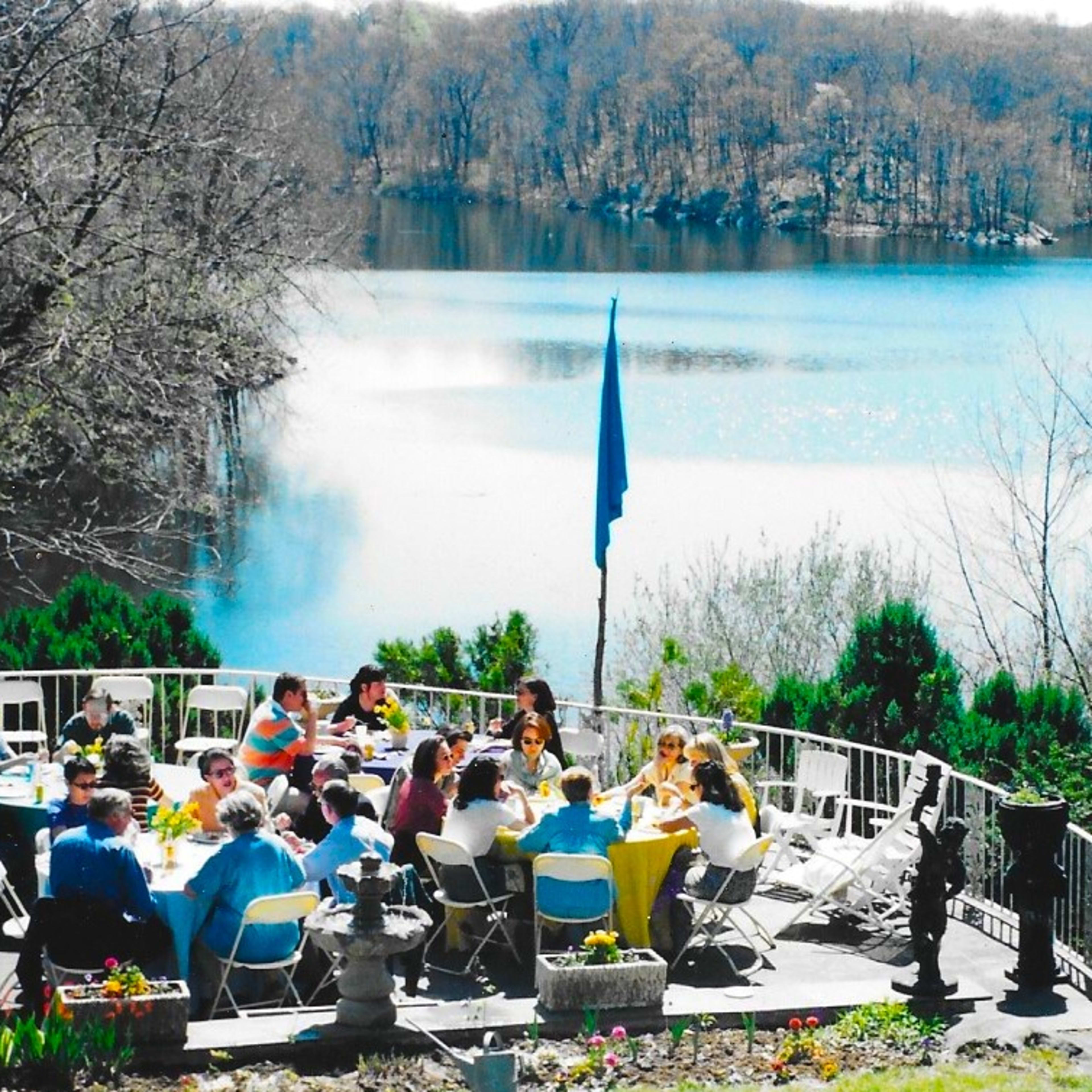 A large group of people sits at tables on a patio overlooking a calm lake surrounded by trees.