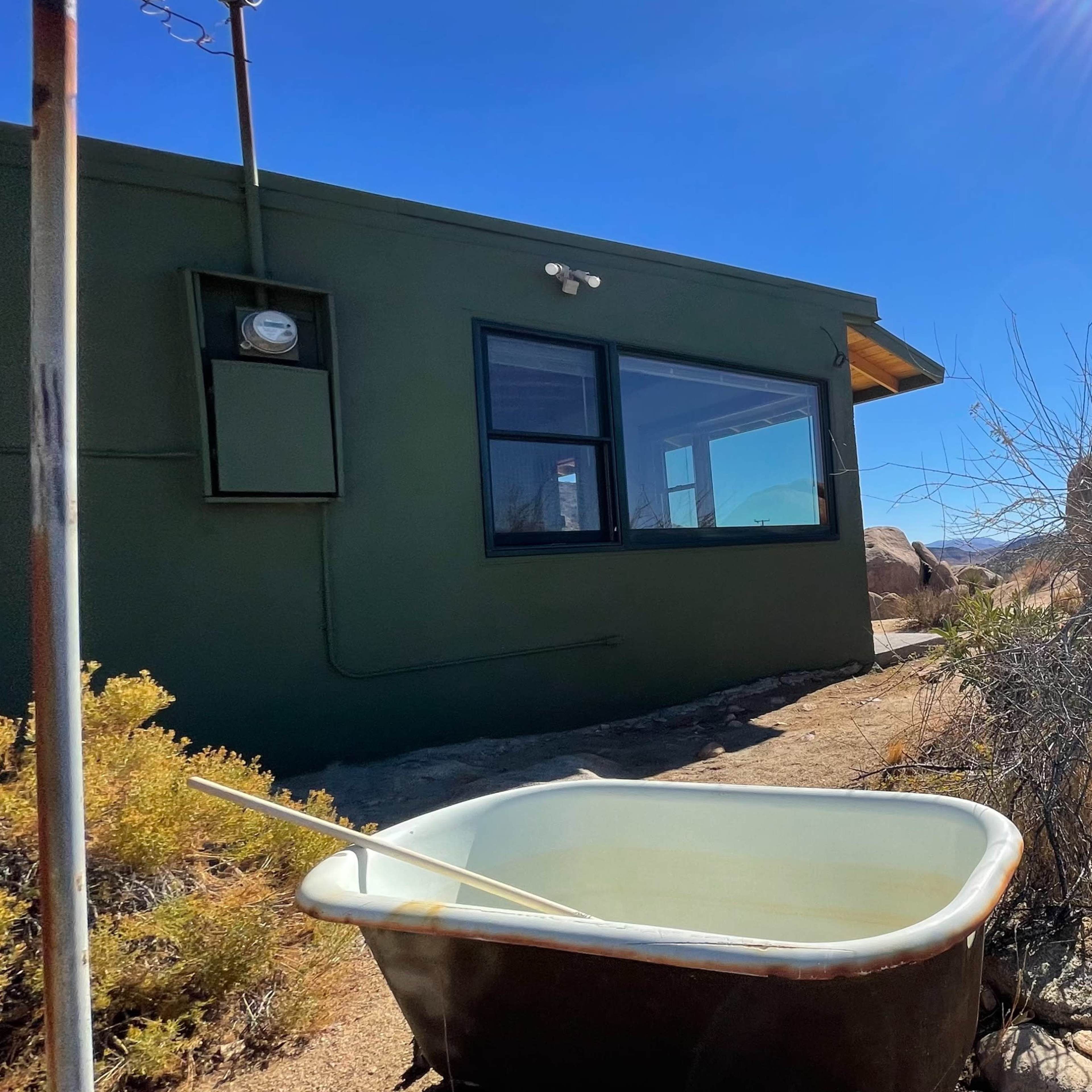 A vintage metal bathtub sits on a rocky pathway beside a green building and boulders under a clear blue sky.