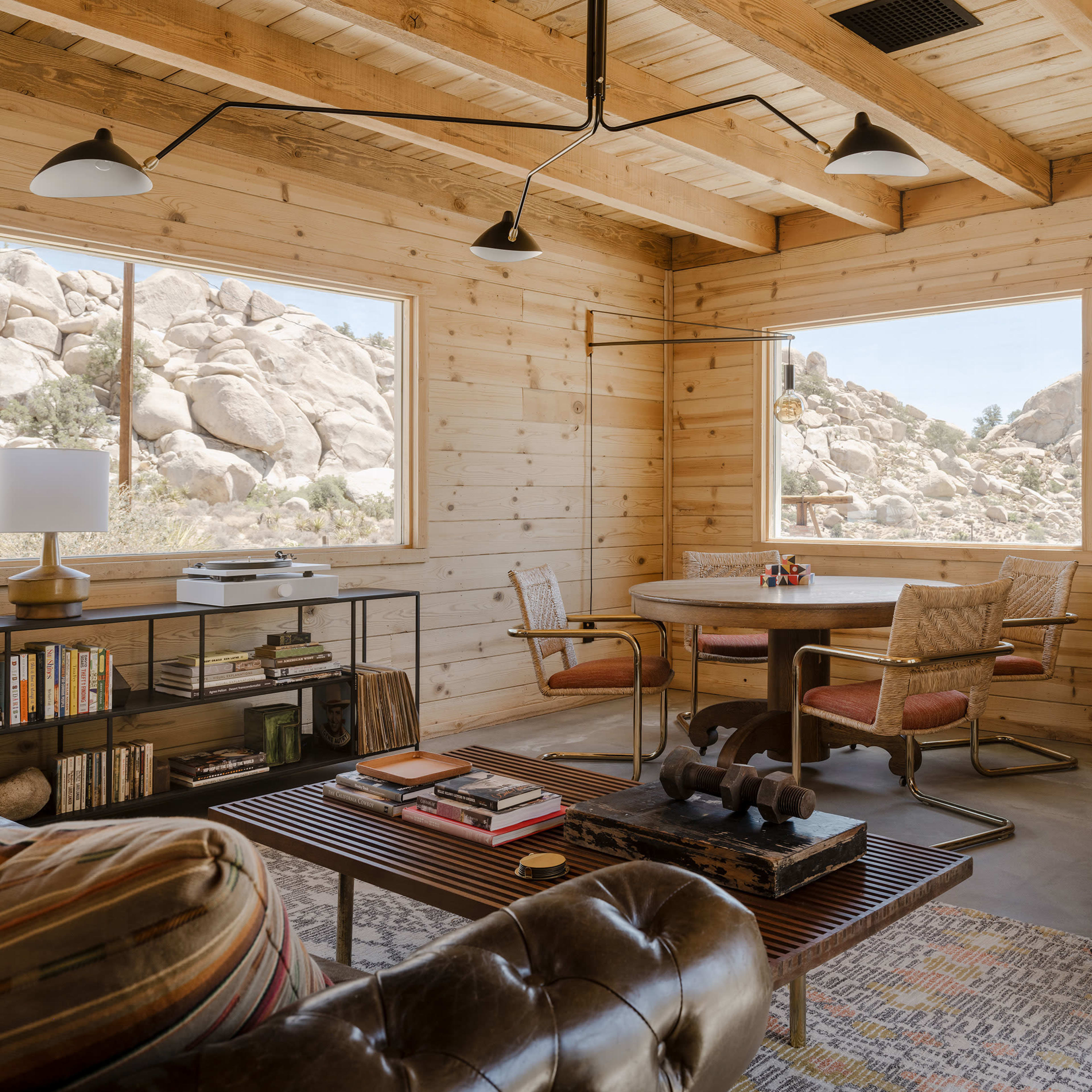 A wooden interior living space with a round table, bookshelf, and a seating area featuring a leather couch and armchairs, all surrounded by large windows that offer a view of rocky terrain outside.