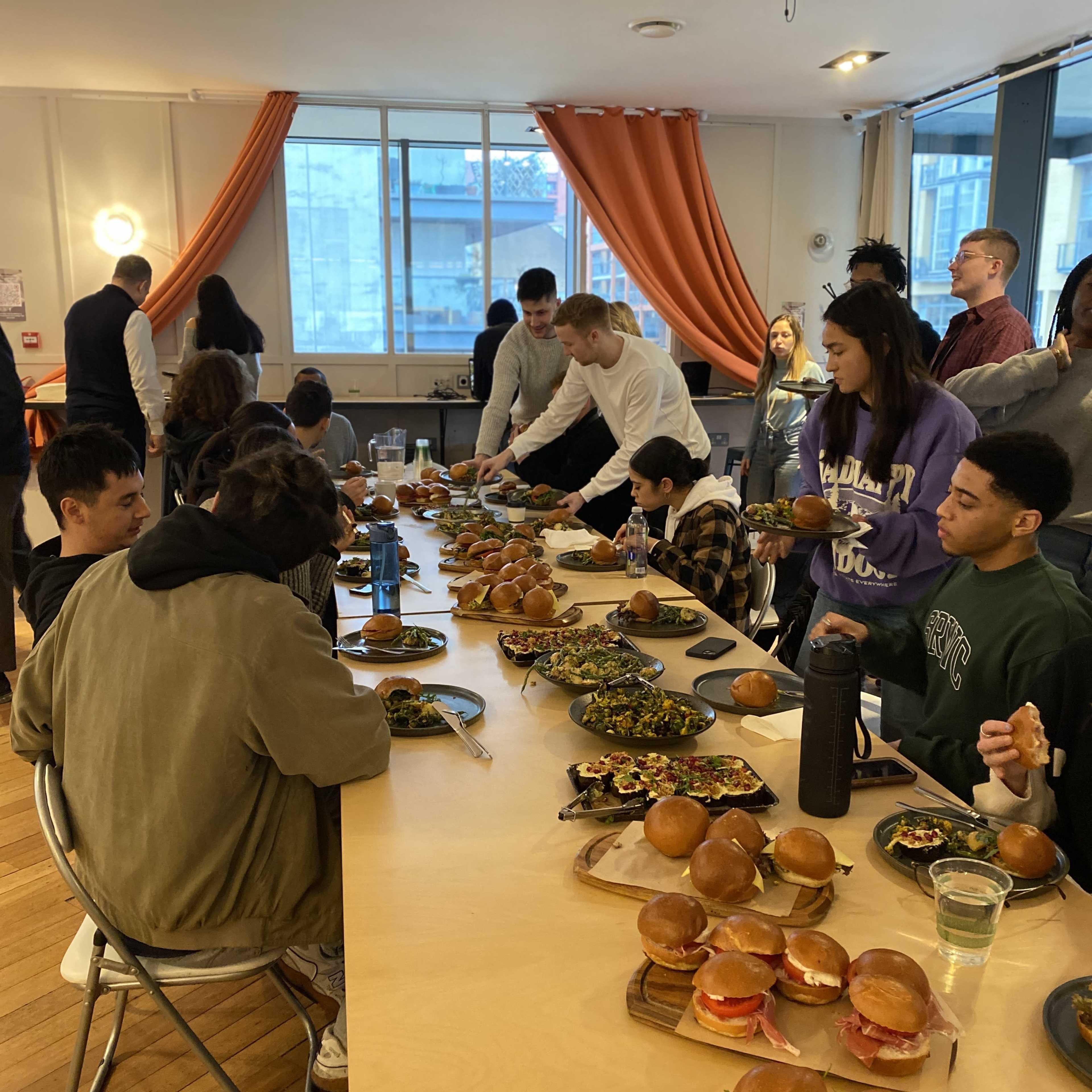 A group of people is gathered around a long table filled with burgers and salads in a well-lit room.