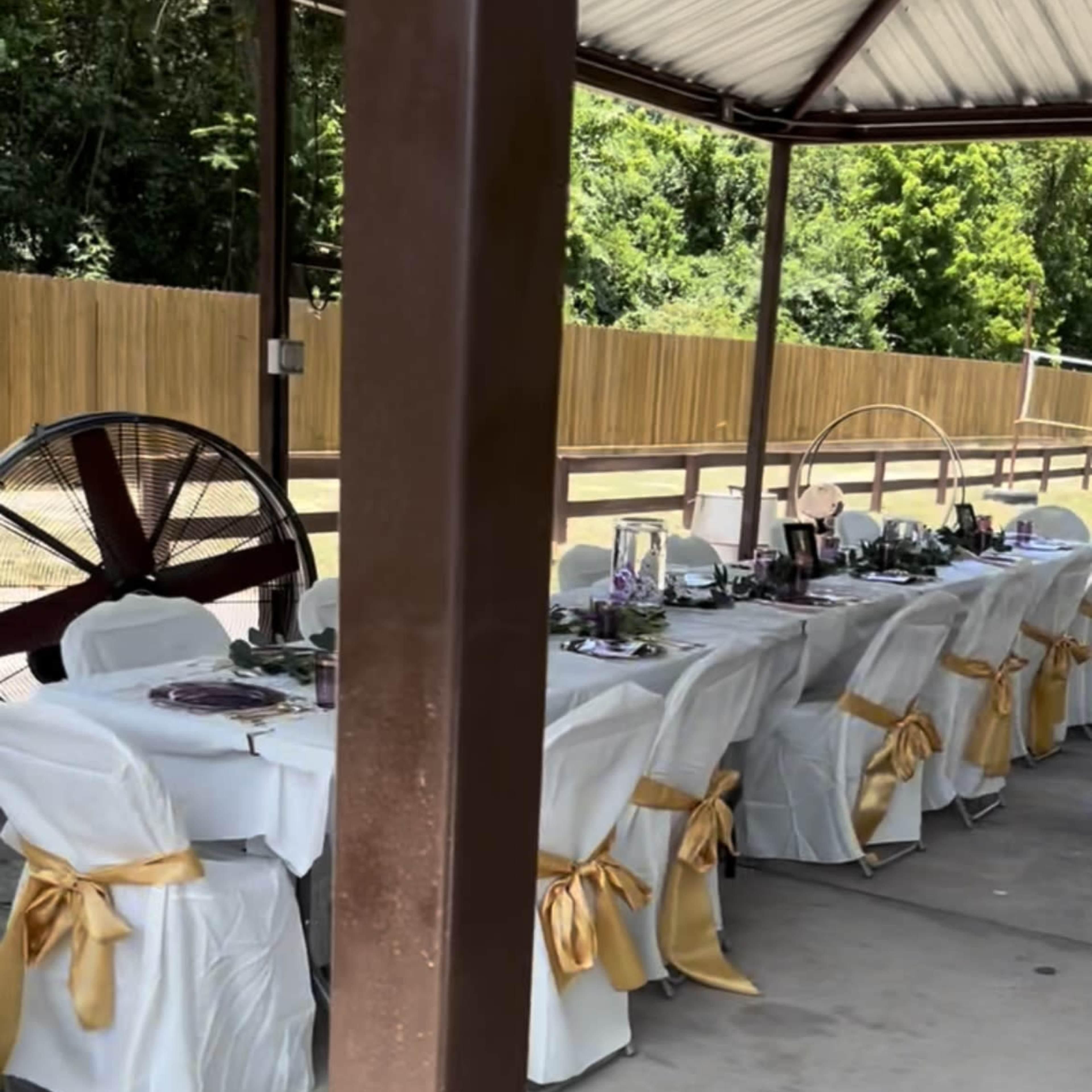 A row of elegantly set dining tables with white tablecloths and gold accents is arranged under a covered patio, surrounded by green trees.