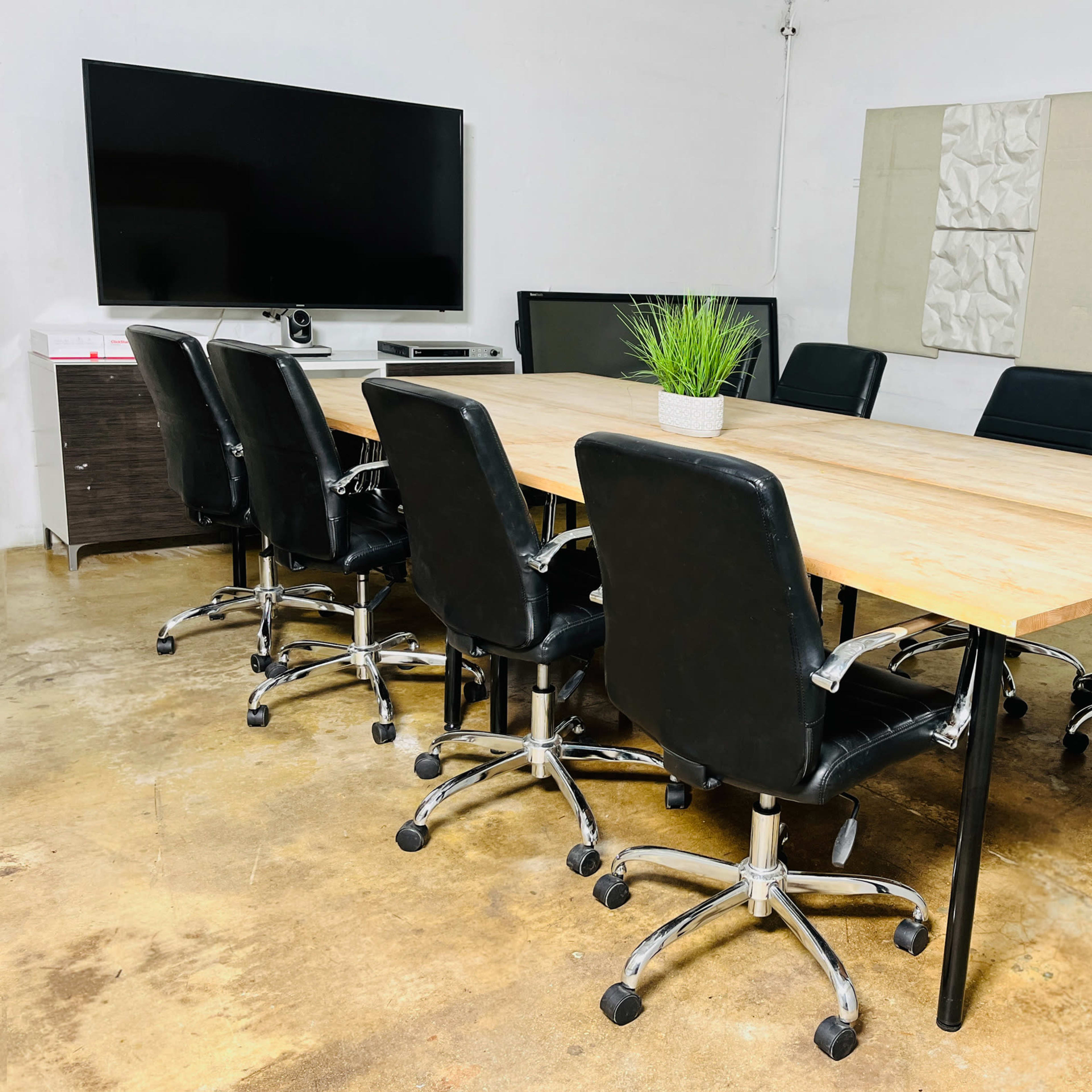 A conference room features a wooden table surrounded by seven black swivel chairs, with a television mounted on the wall and a small plant on the table.