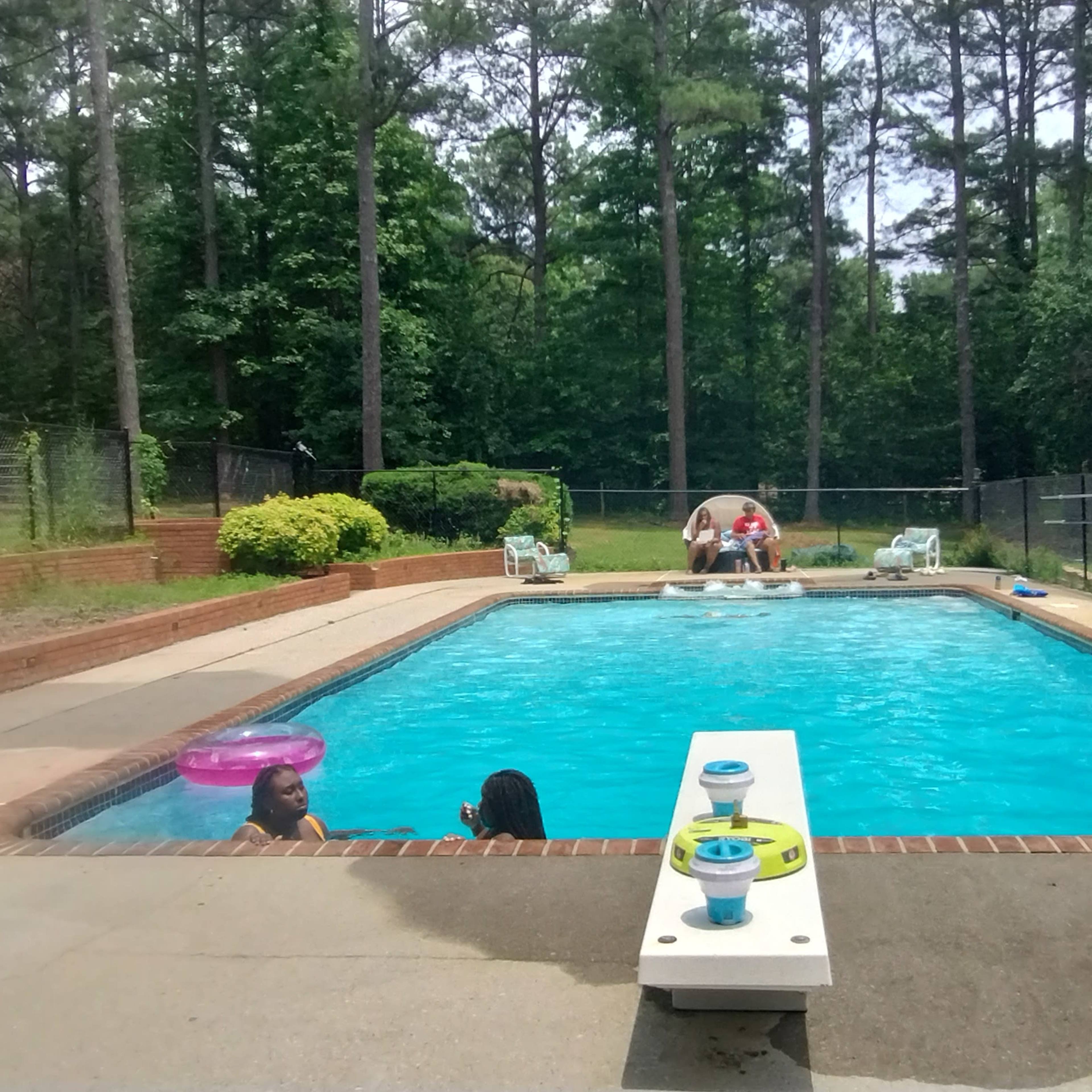 A backyard swimming pool surrounded by trees, with several people relaxing in and beside the water.