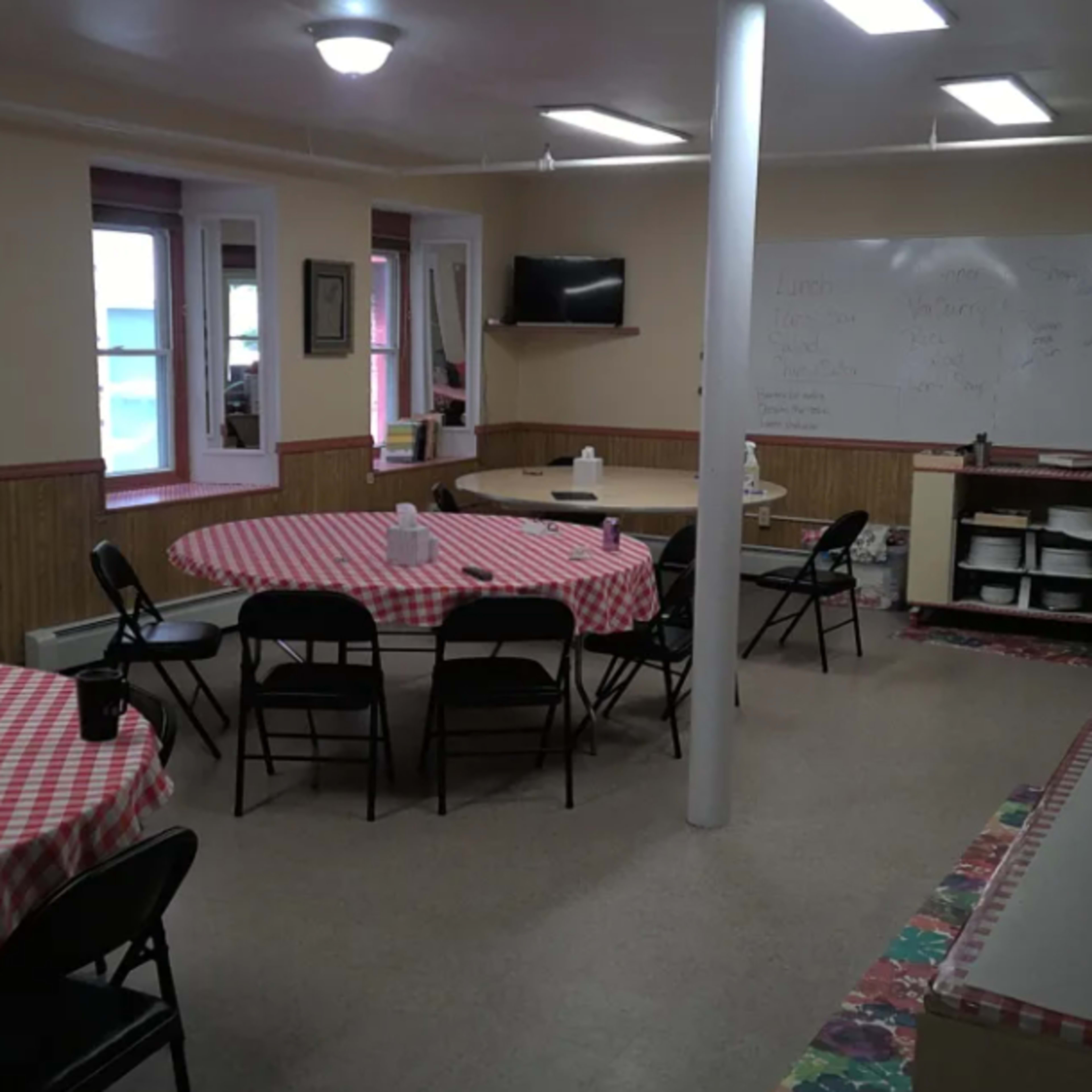 A spacious indoor dining area with several round tables covered in red and white checkered tablecloths, a kitchenette along one wall, and a whiteboard on another.