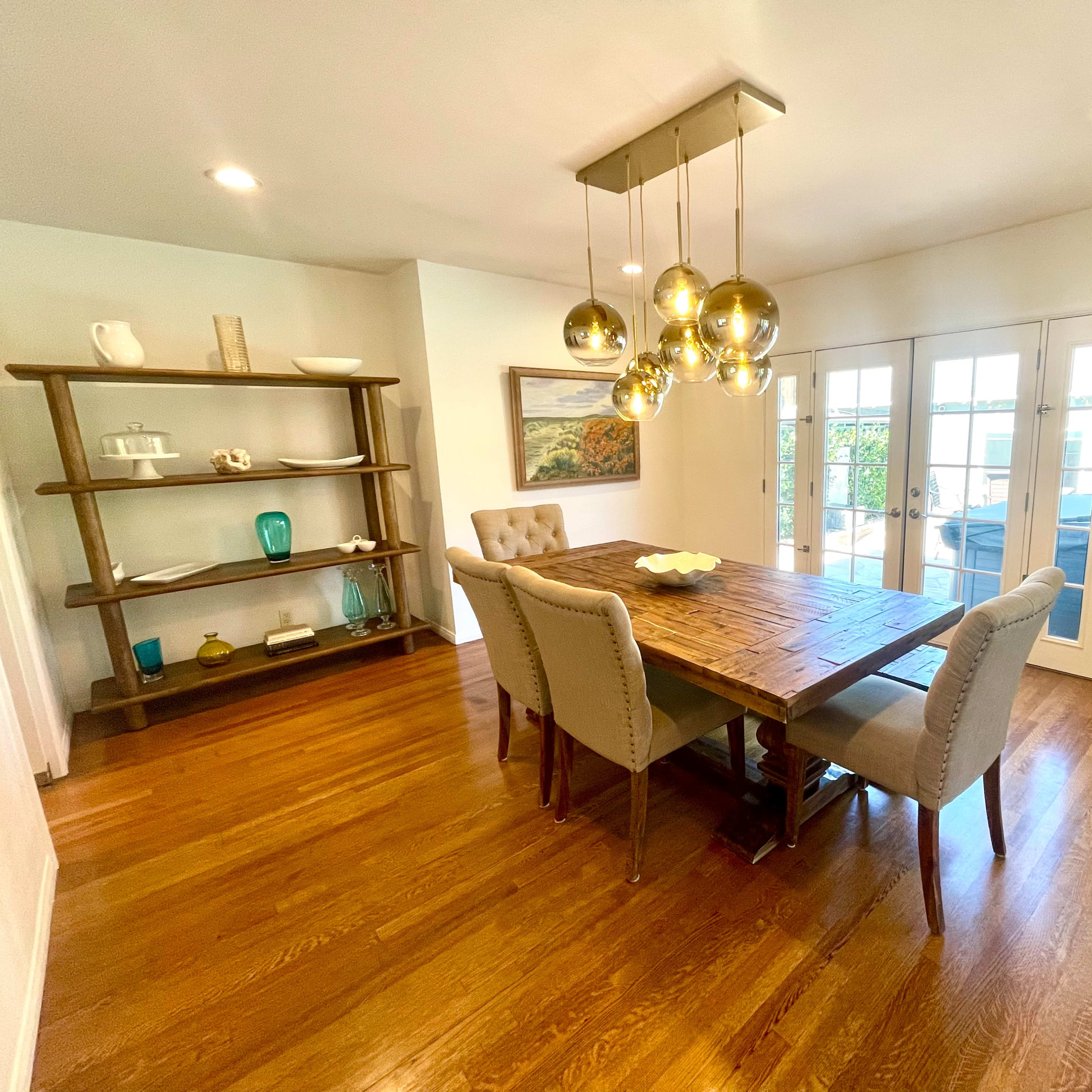 A dining room features a wooden table with upholstered chairs, a shelf displaying decorative items, and a light fixture above.