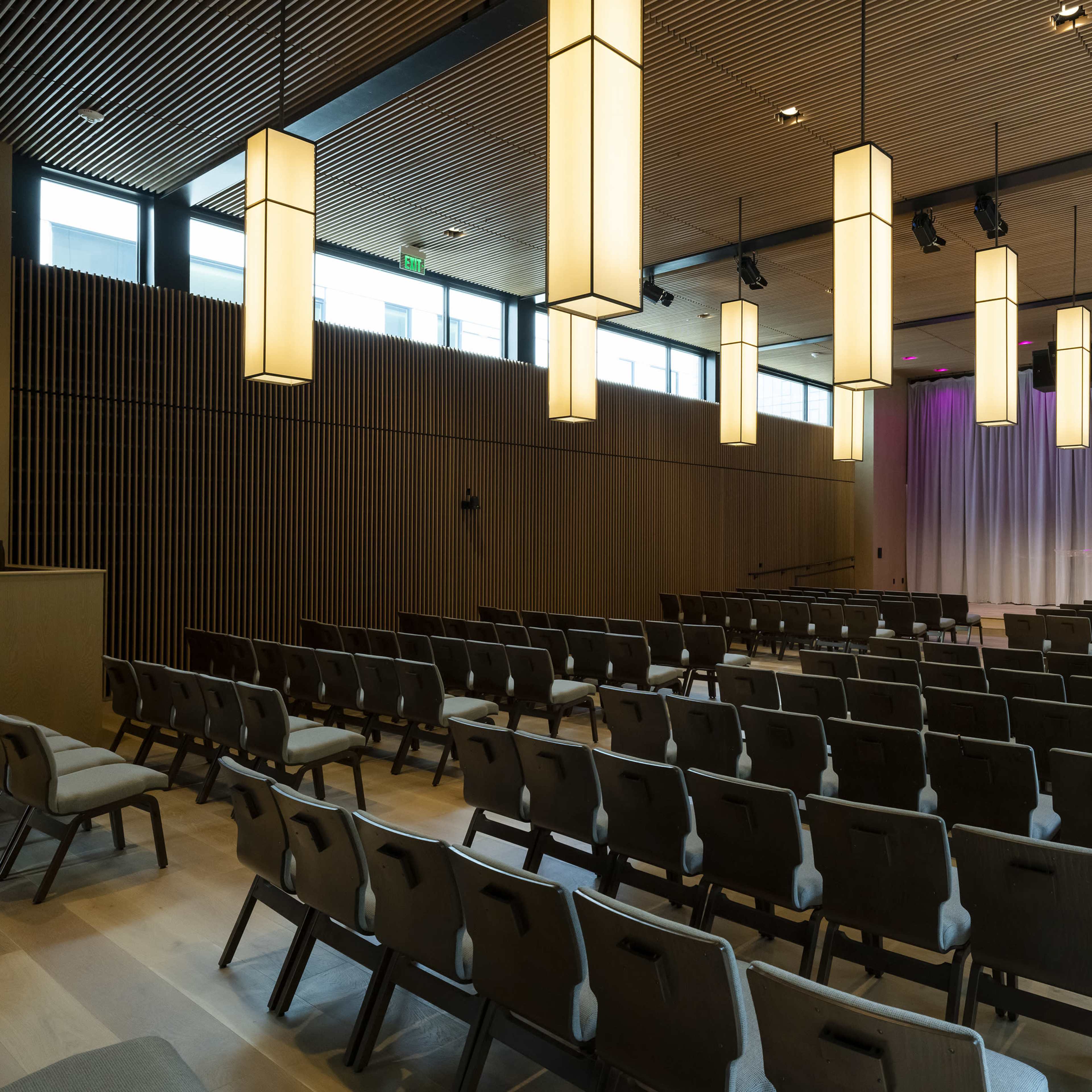 A modern auditorium with rows of chairs facing a stage, accented by hanging light fixtures and large windows.