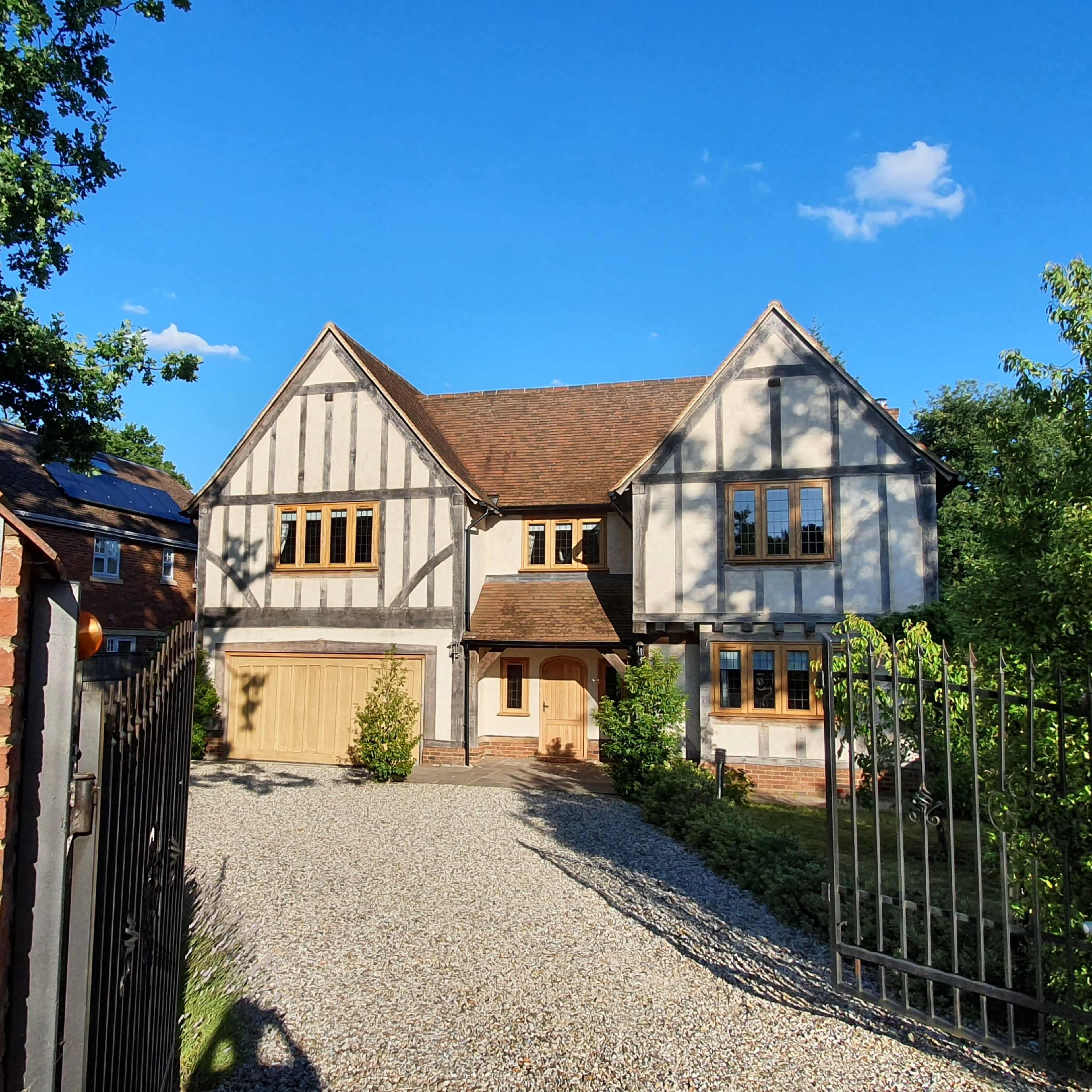 A two-story house with a half-timbered design and a gravel driveway is surrounded by greenery.