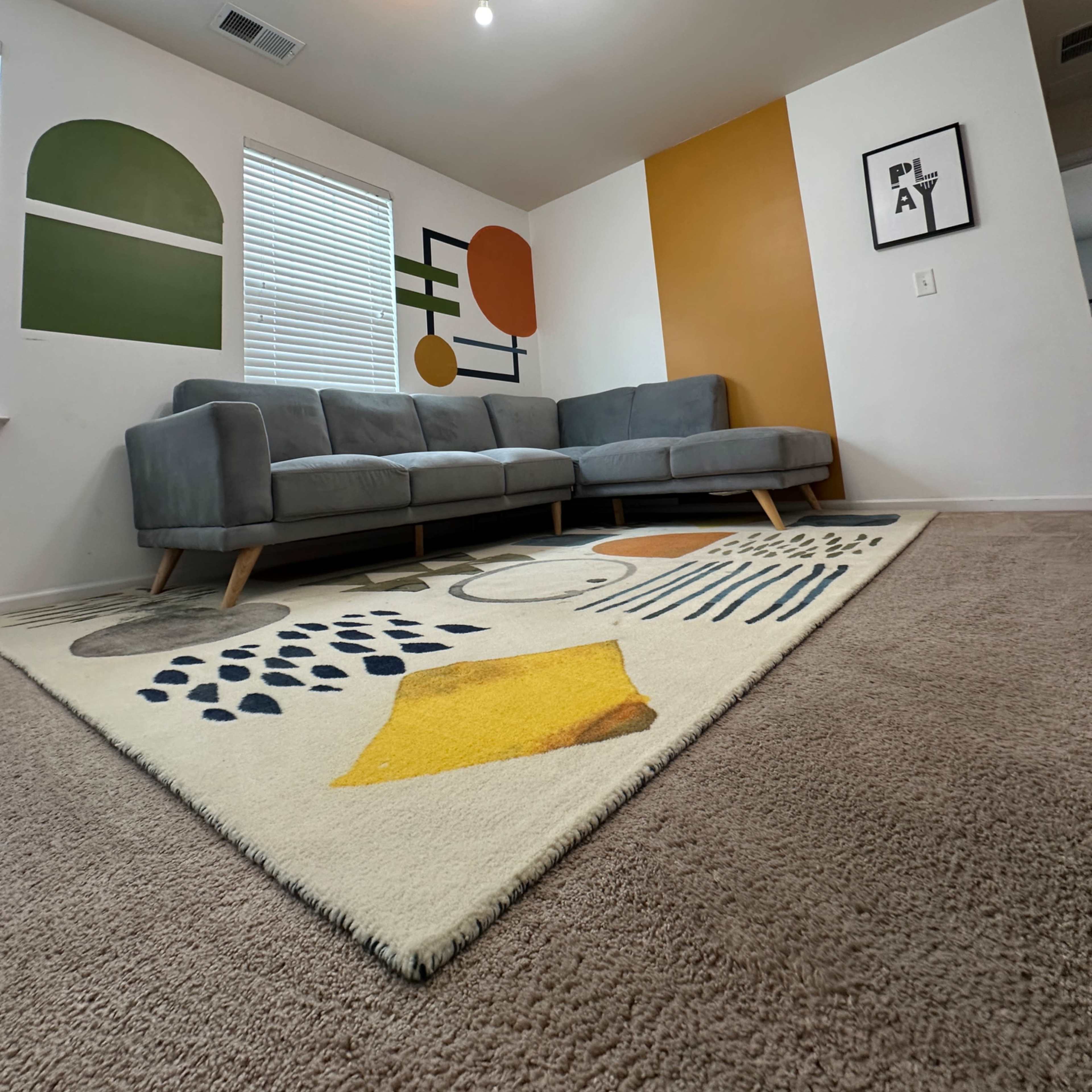 A gray sectional sofa is positioned in a brightly lit room featuring abstract wall art and a large patterned area rug.