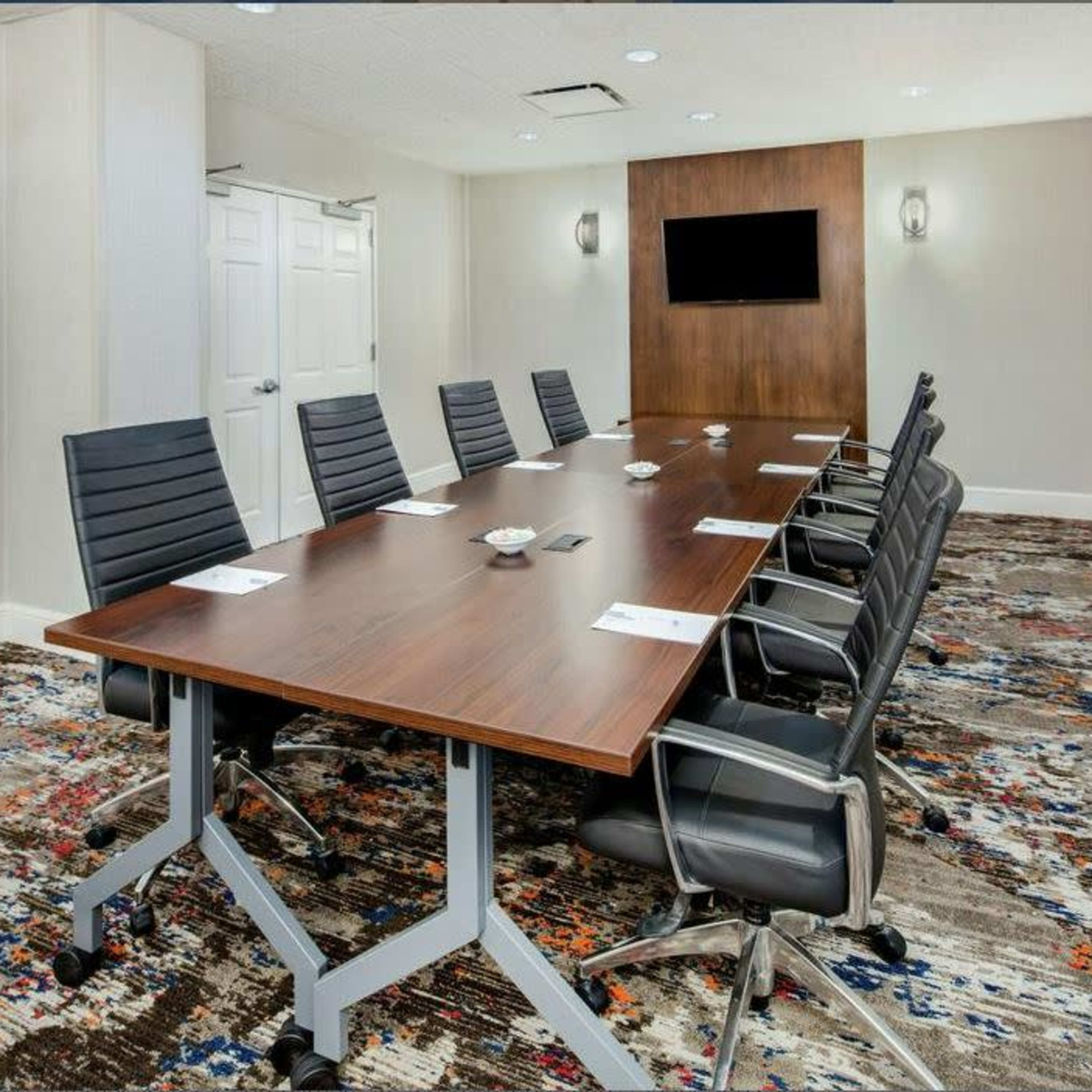 A modern conference room features a long wooden table surrounded by black office chairs, with a wall-mounted screen and double doors at either end.
