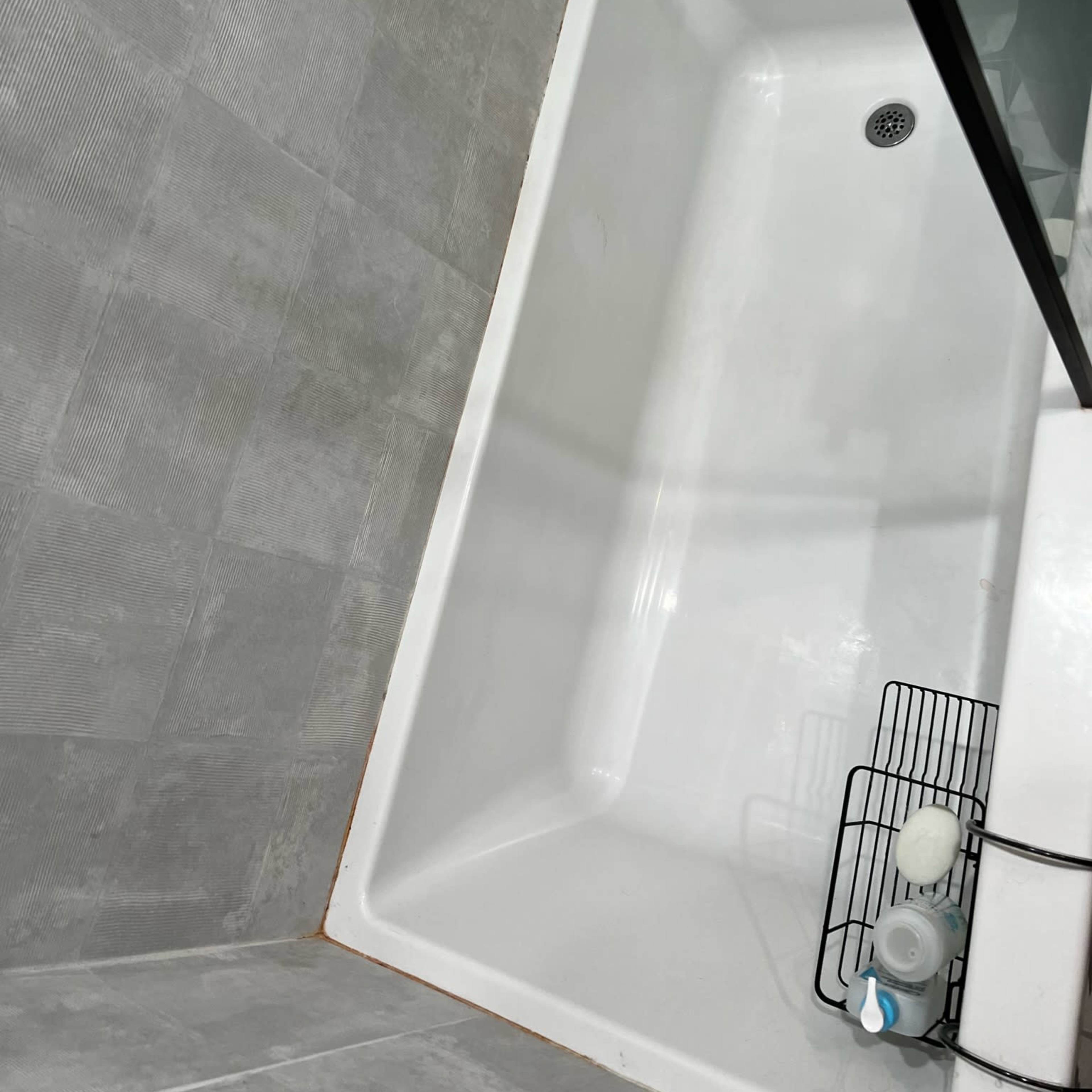 A top-down view of a white bathtub with a glass shower screen and a black metal shower caddy containing bath products.