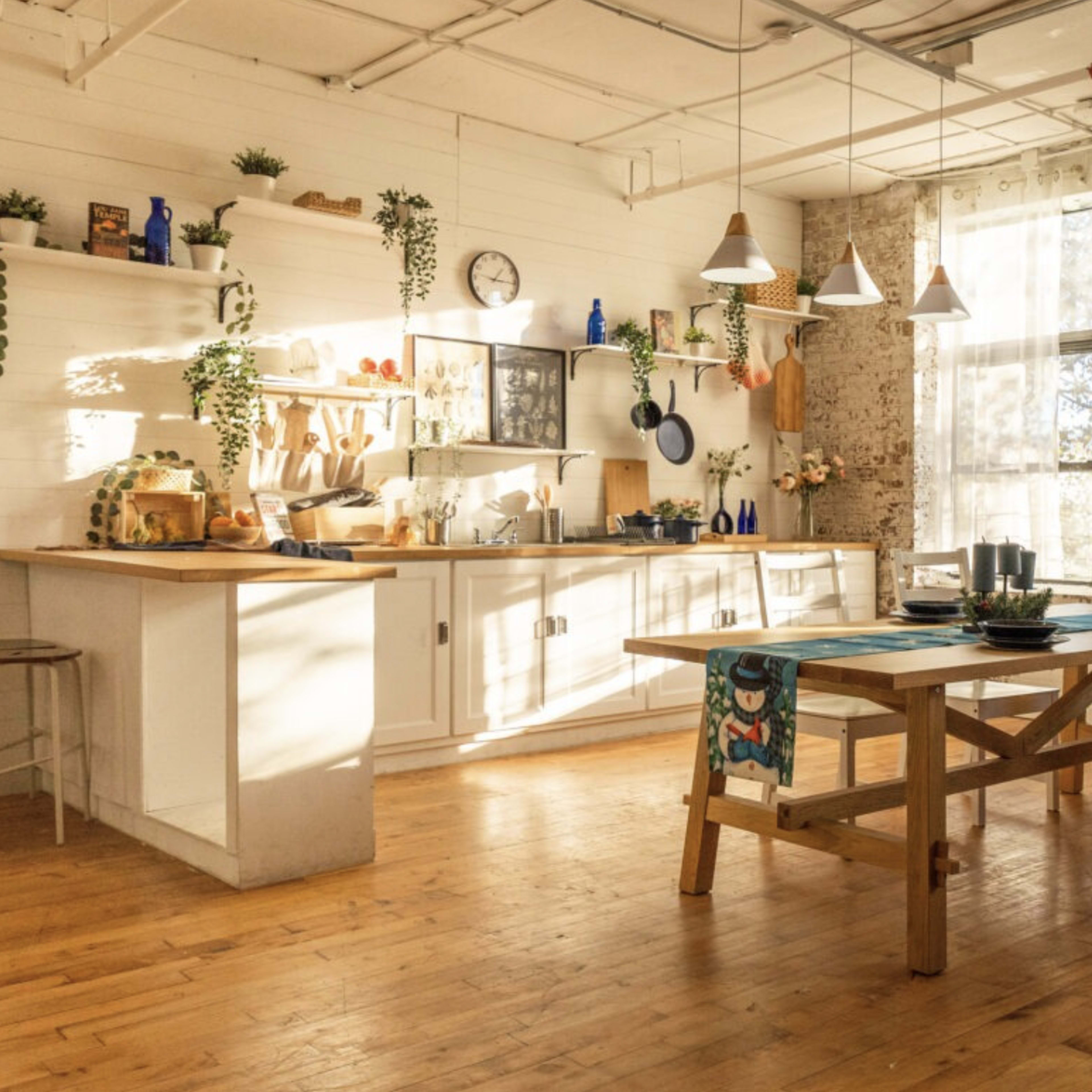 A bright, open kitchen with wooden floors, a dining table, and shelves filled with various kitchen items, decorated with plants and warm sunlight streaming through the windows.