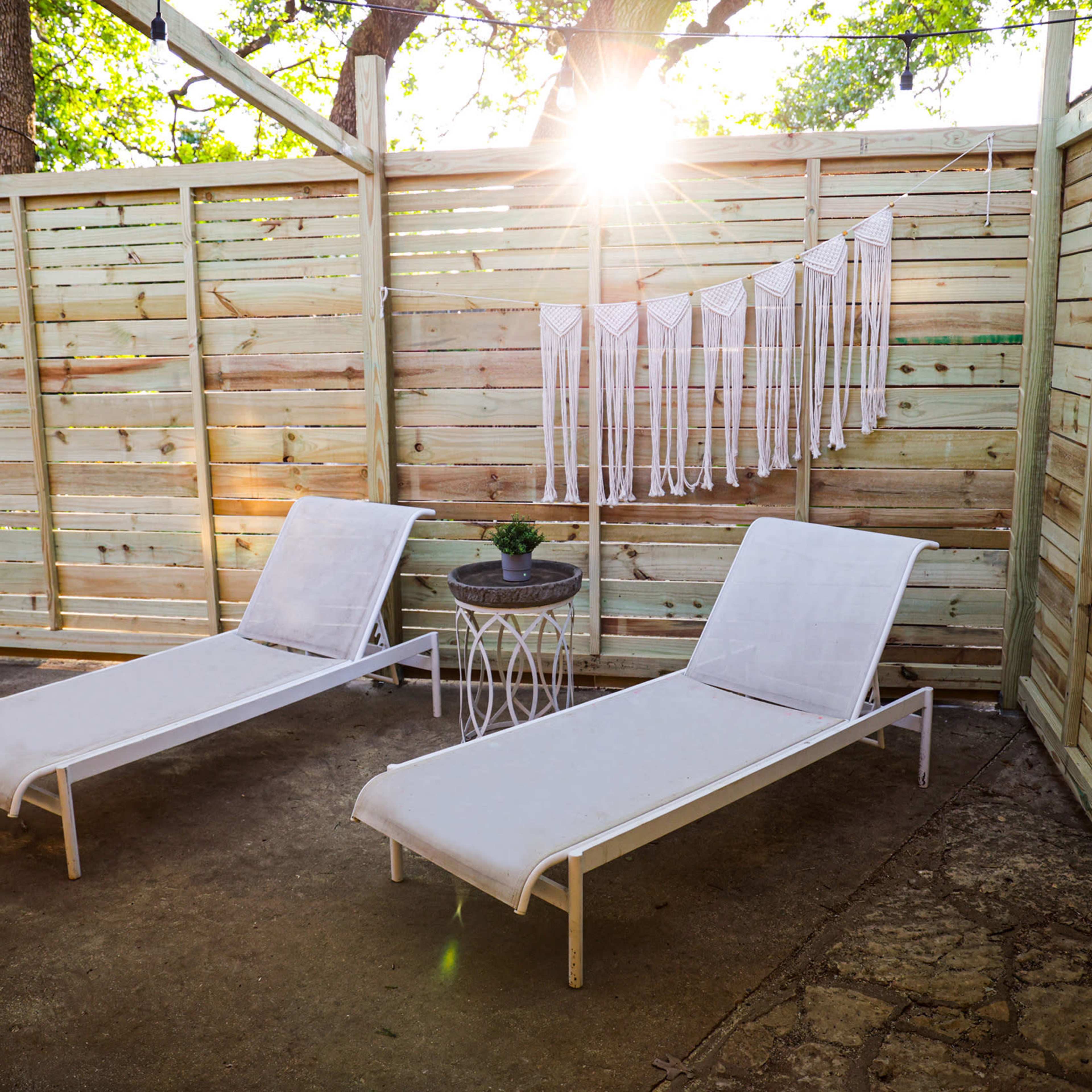 Two white lounge chairs positioned beside a small table with a plant, under a wooden structure with a sunbeam shining through the trees.