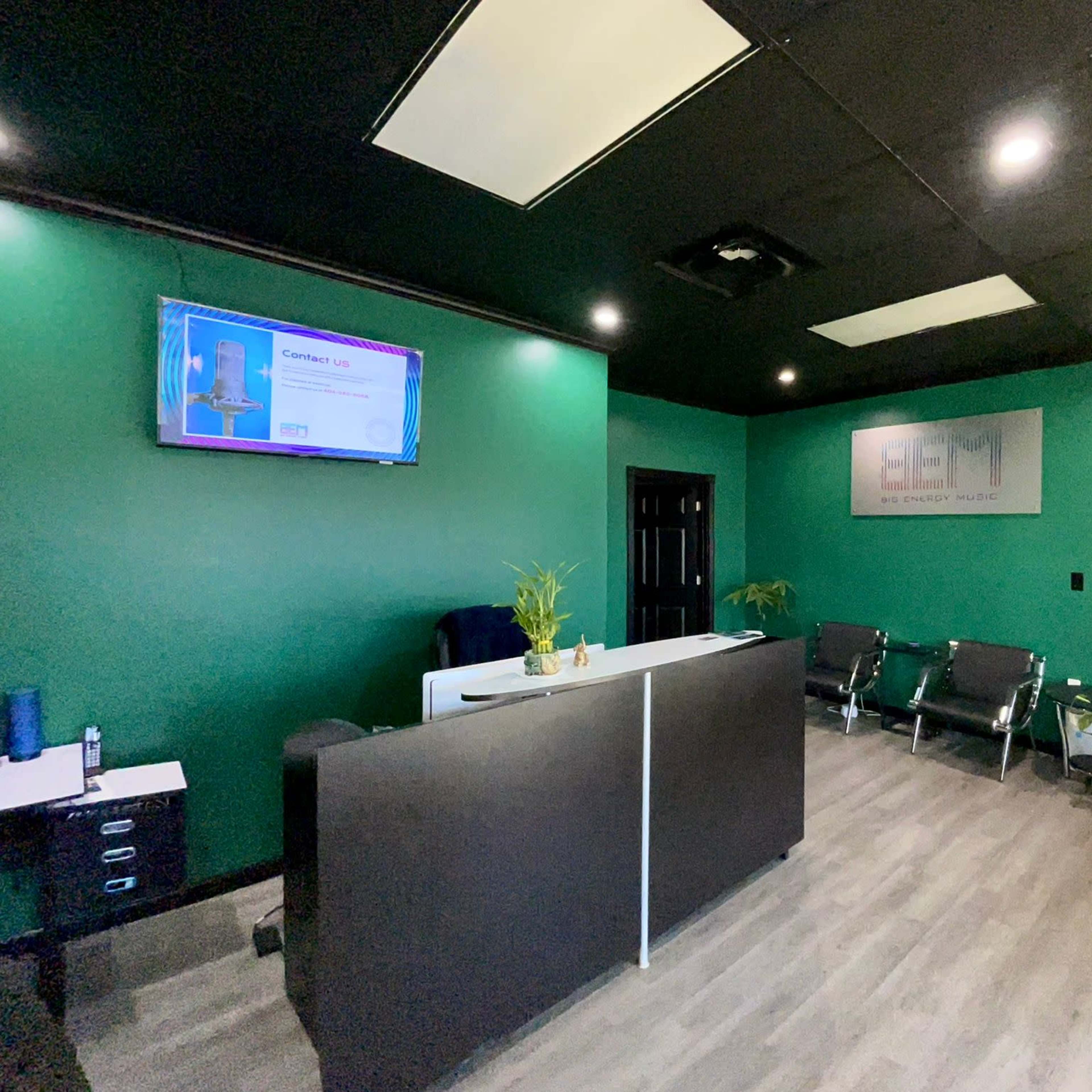 A small reception area with a dark green wall, a front desk, a television displaying information, and several chairs arranged for visitors.