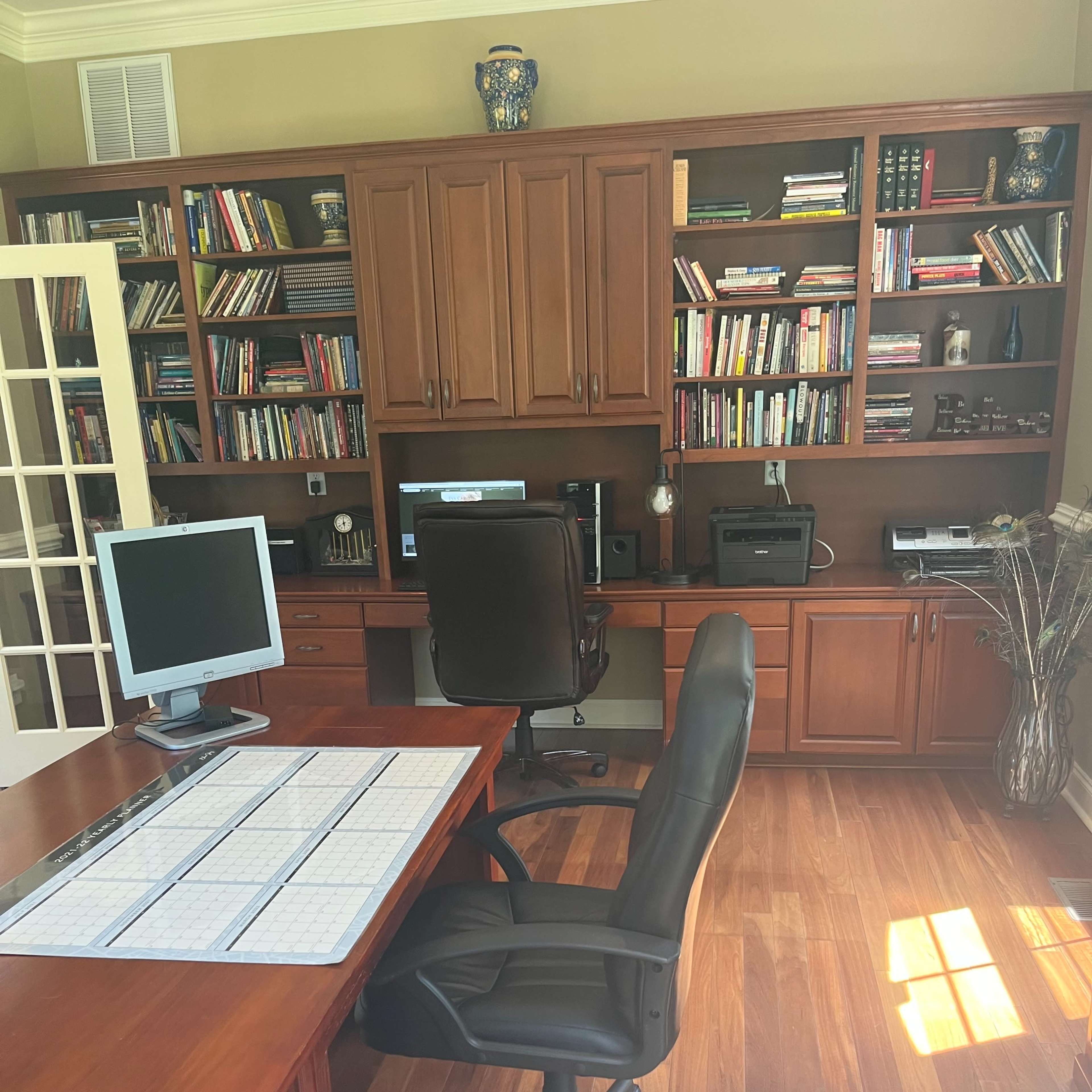 A home office with a wooden desk, a swivel chair, two monitors, and a bookshelf filled with books against a wall.