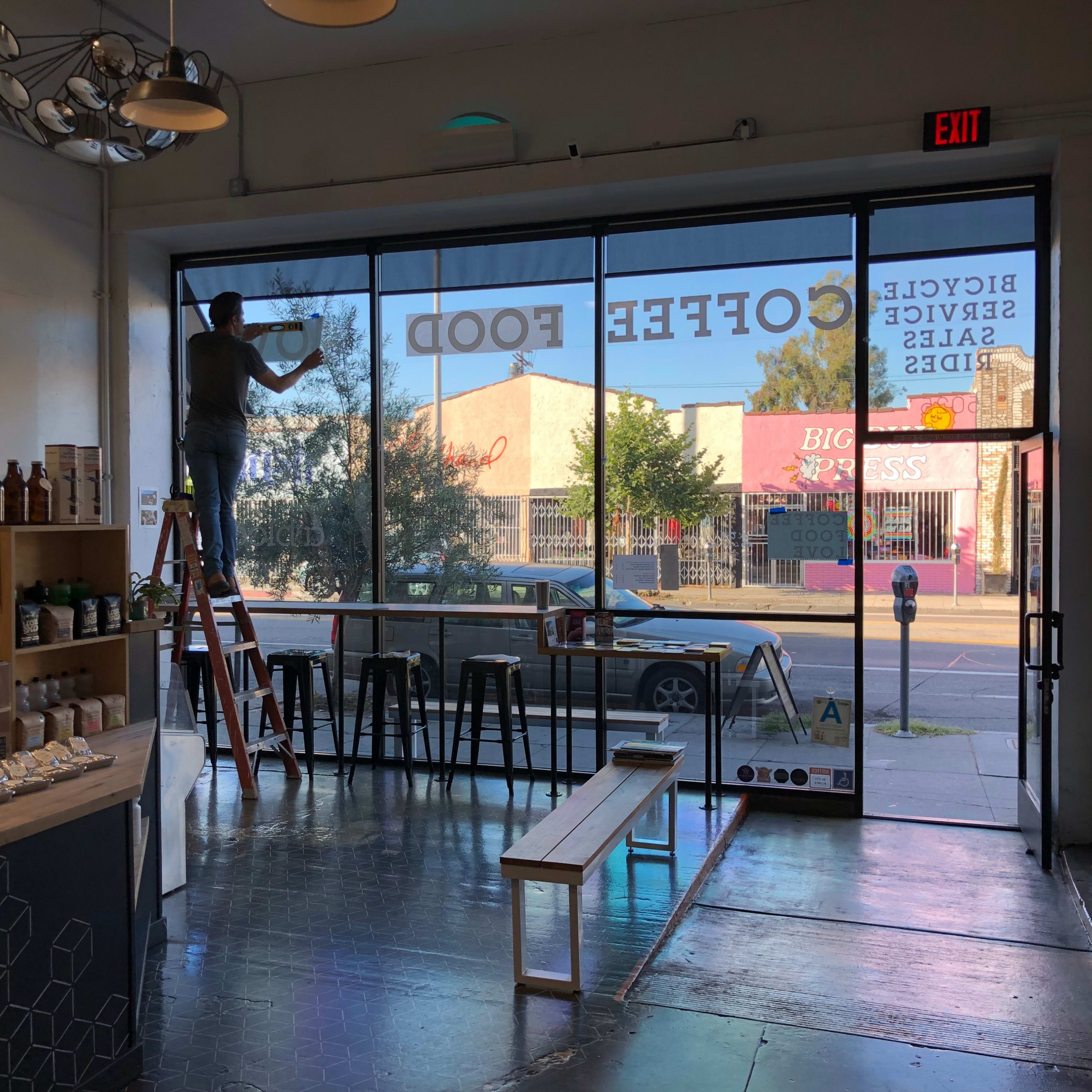 A person is standing on a ladder applying a sign to the window of a coffee shop.