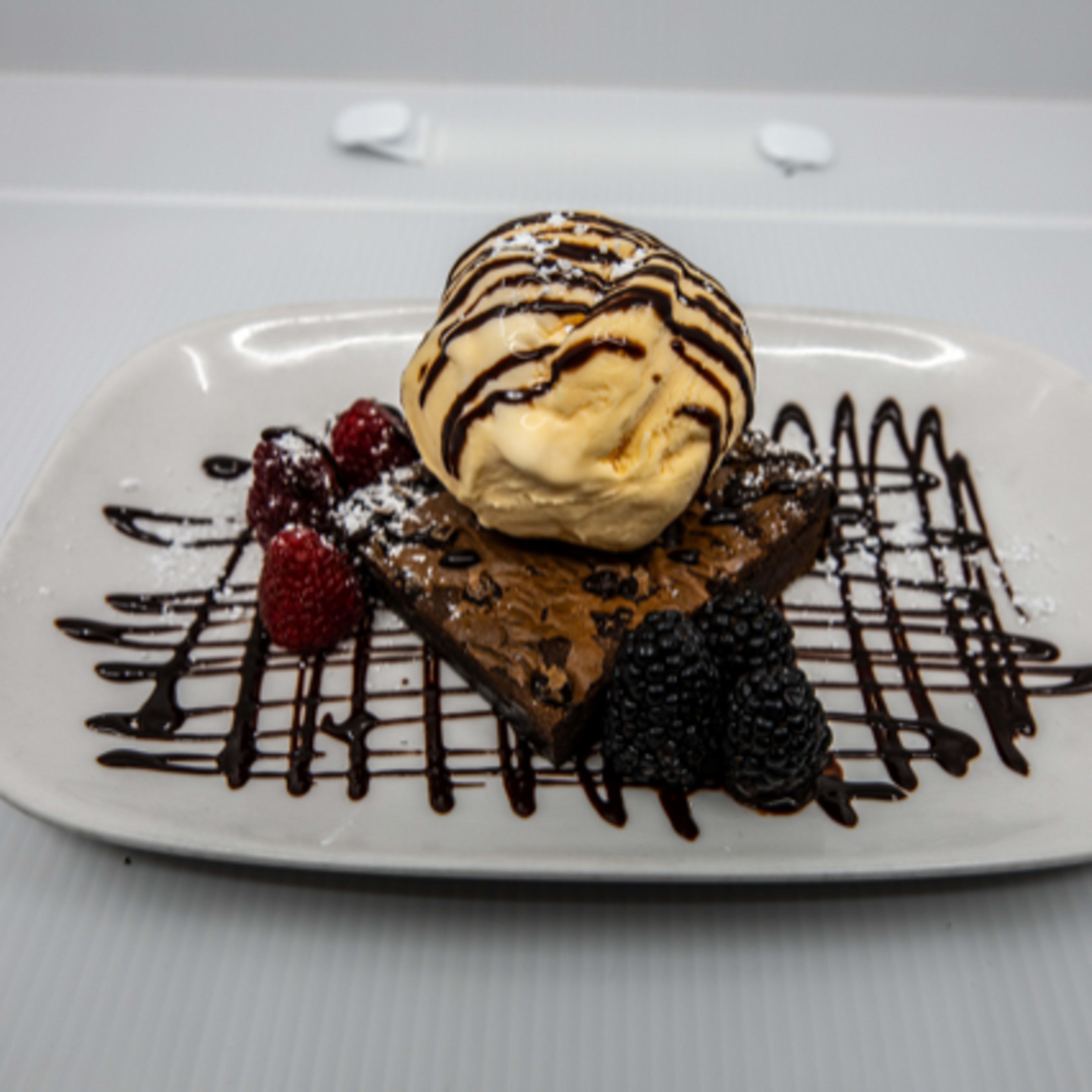 A rectangular brownie topped with a scoop of vanilla ice cream, drizzled with chocolate sauce, and garnished with raspberries and blackberries on a white plate.
