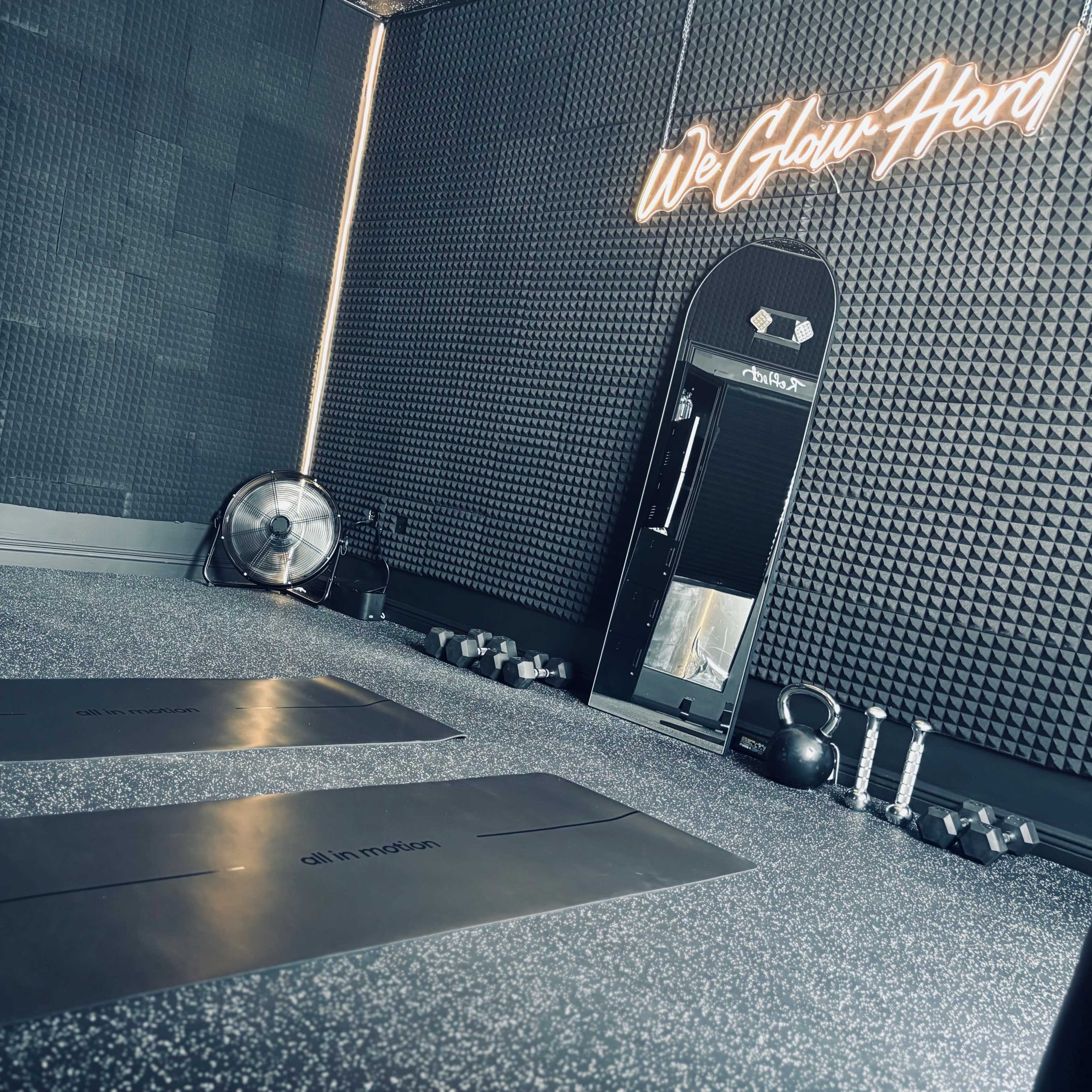 A modern gym interior with two exercise mats, kettlebells, water bottles, and a fan against a textured wall featuring neon signage.