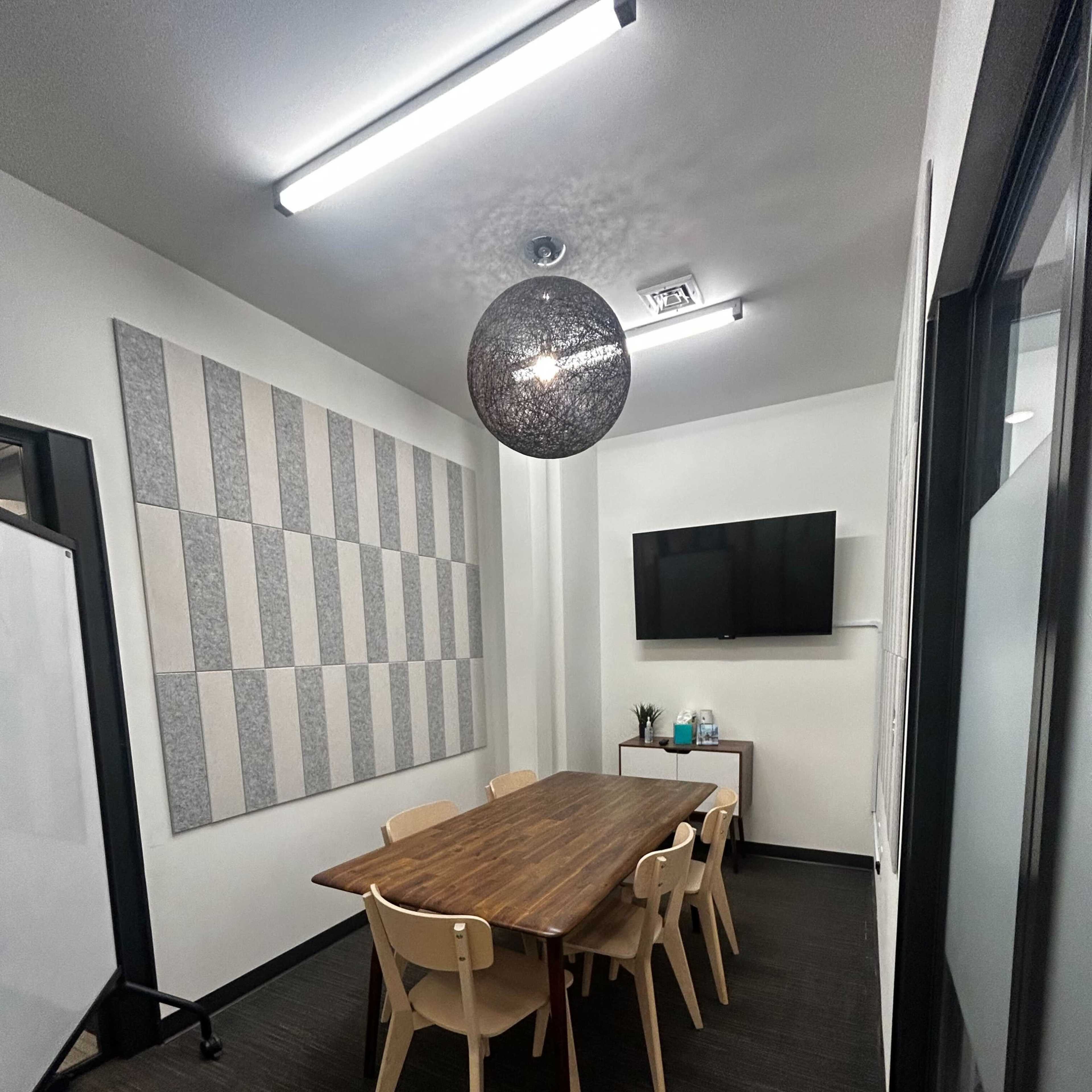 A conference room with a large wooden table surrounded by six chairs, a television mounted on the wall, and acoustic panels on one side.