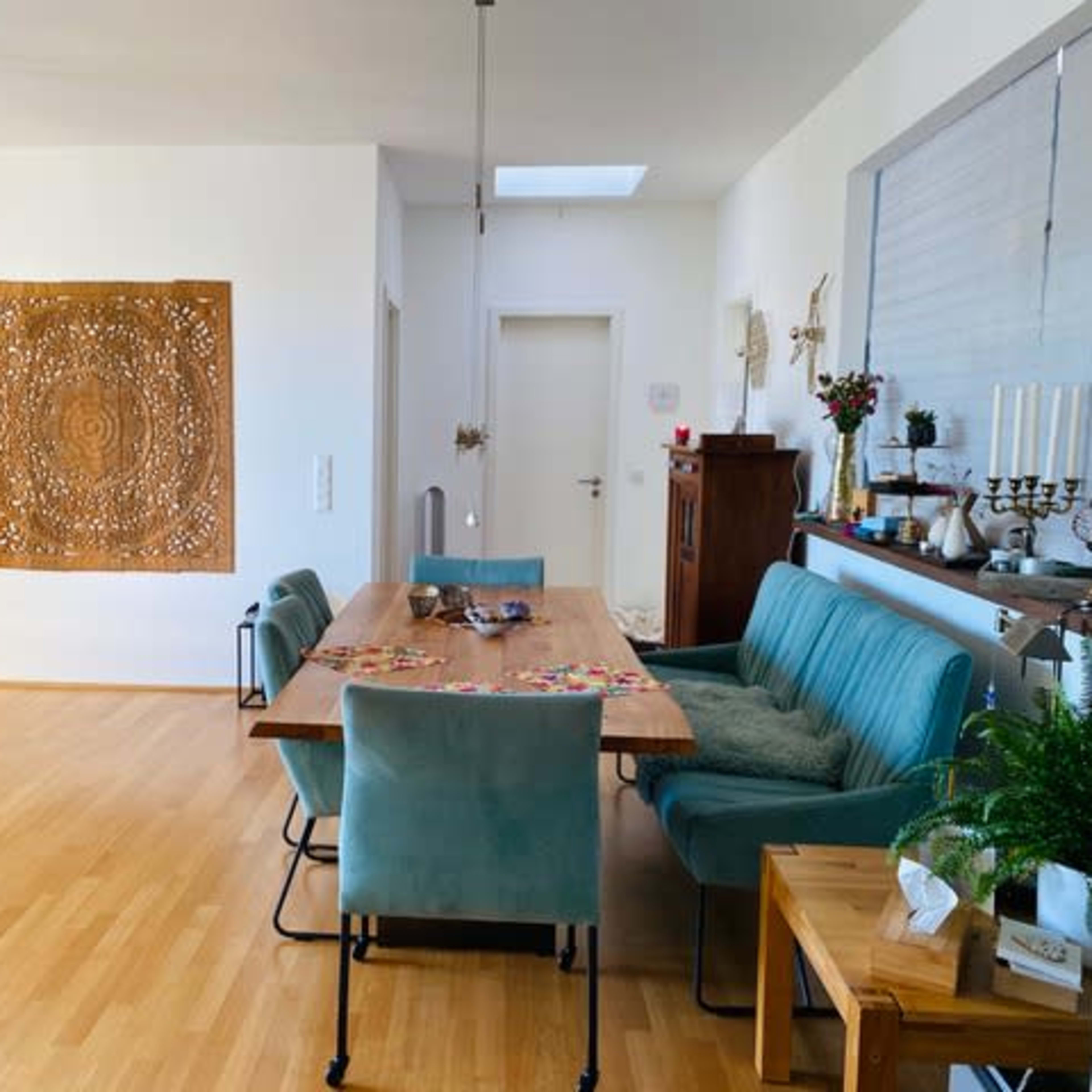 A spacious dining area features a wooden table surrounded by teal chairs, with a decorative wall hanging and a dog lying on the floor.