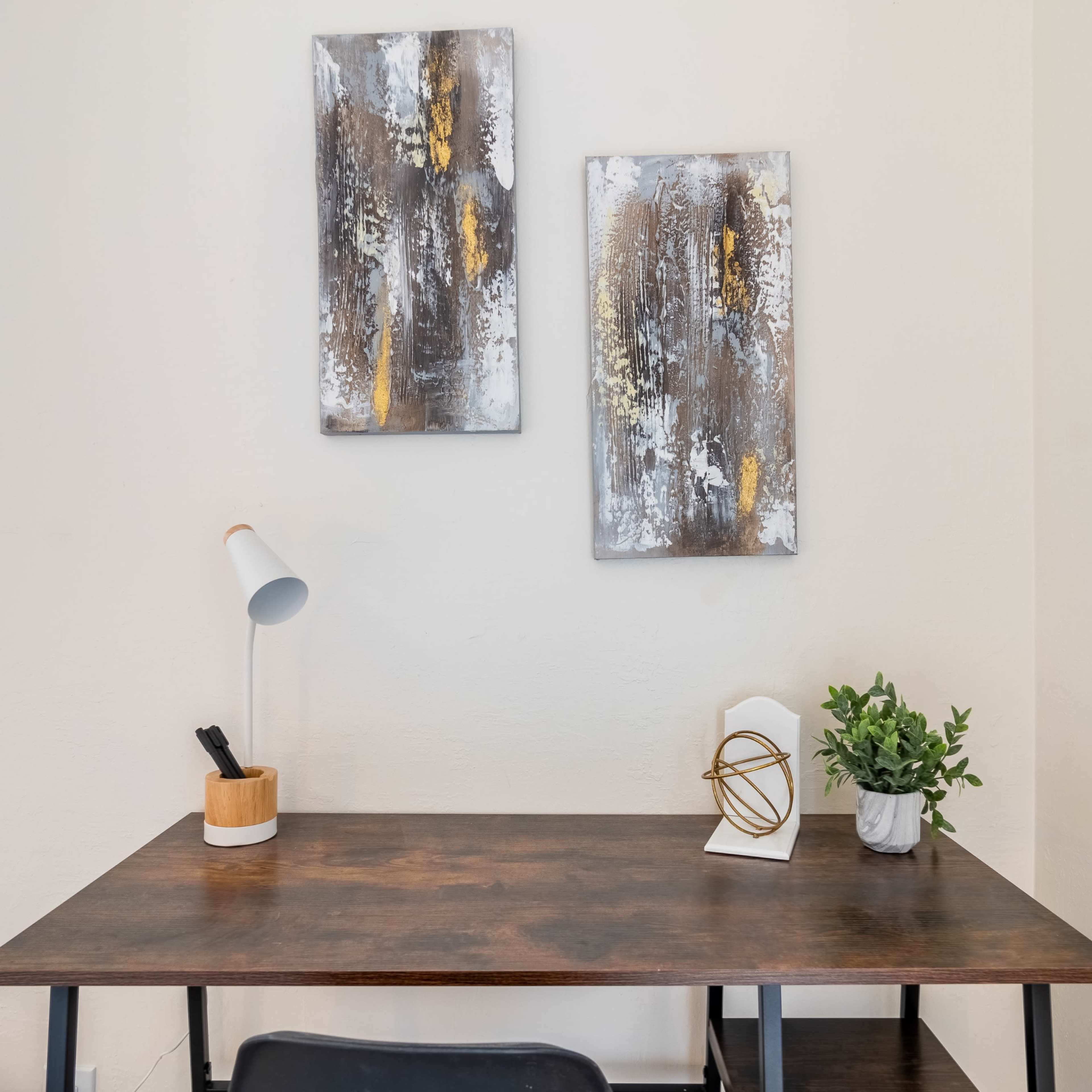 A wooden desk is positioned against a wall, adorned with two abstract paintings above it, a small potted plant, and a desk lamp on the surface.
