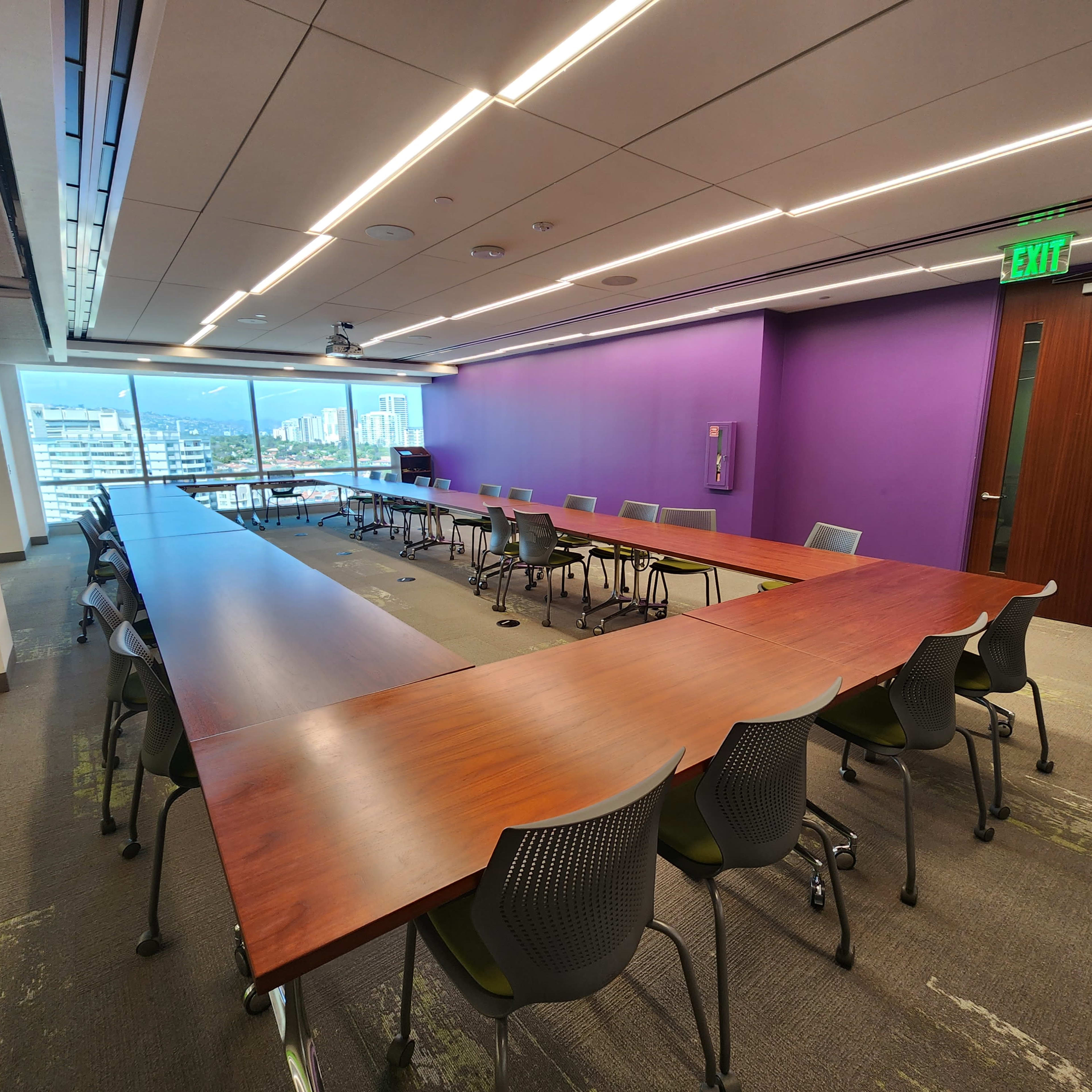 A modern conference room features a large rectangular table surrounded by ergonomic chairs, with a purple accent wall and large windows offering a view of the outside.