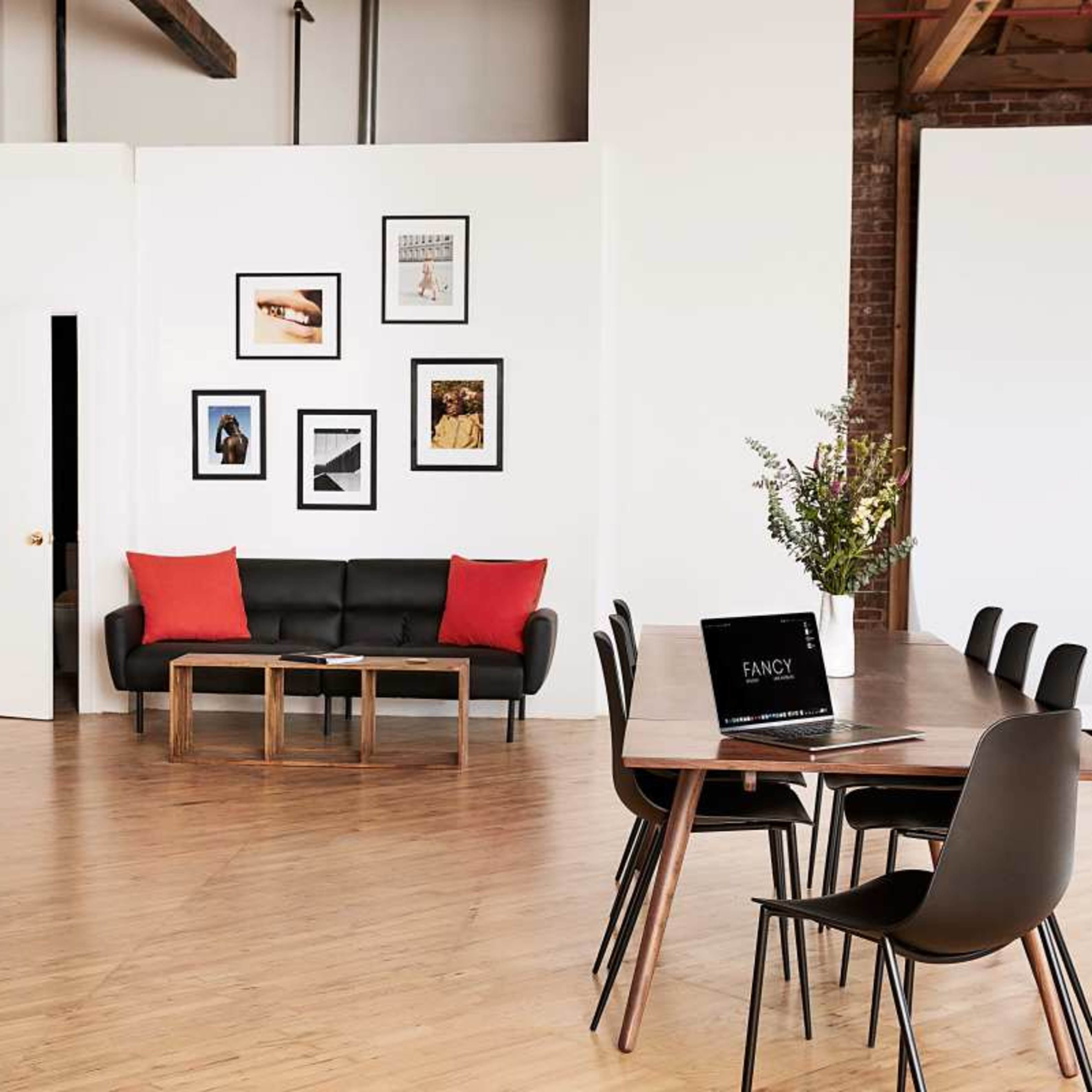 The scene features a modern, open workspace with a black sofa, a wooden coffee table, framed art on the walls, and a large table set for meetings.