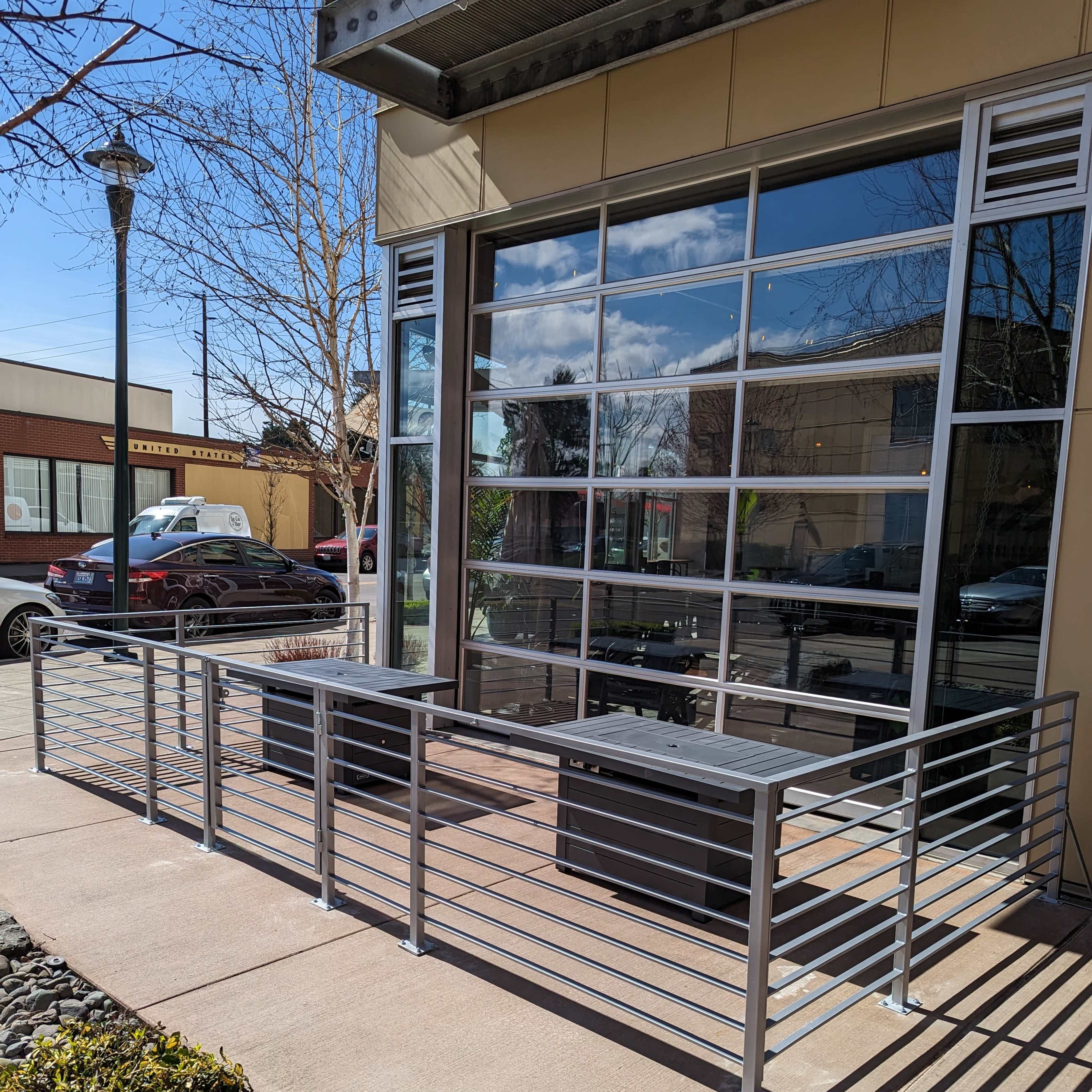 A metal railing enclosing a small outdoor seating area in front of a building with large glass windows.