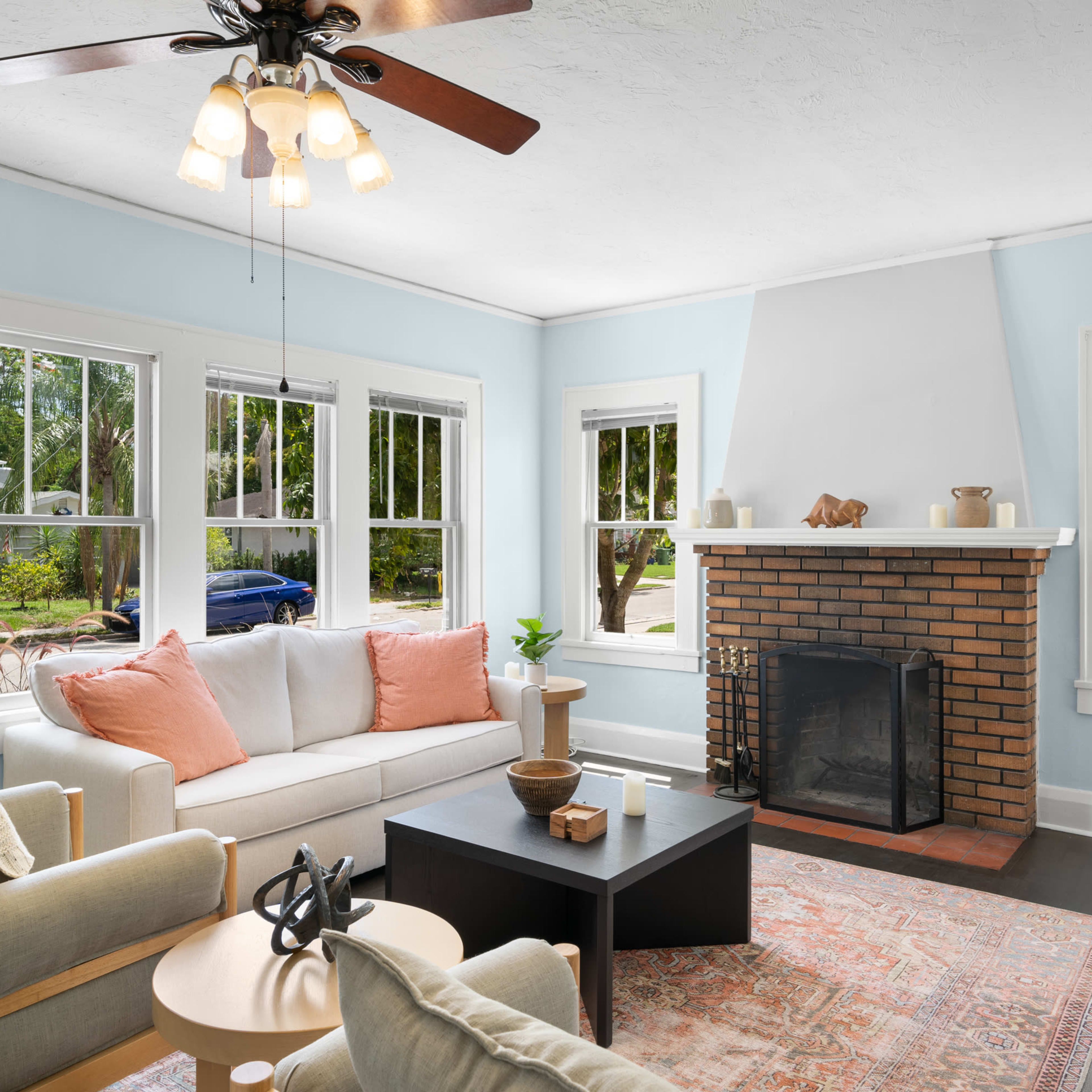 A cozy living room with a light blue wall, a brick fireplace, large windows, and a mix of furniture including a sofa with orange cushions and a black coffee table.