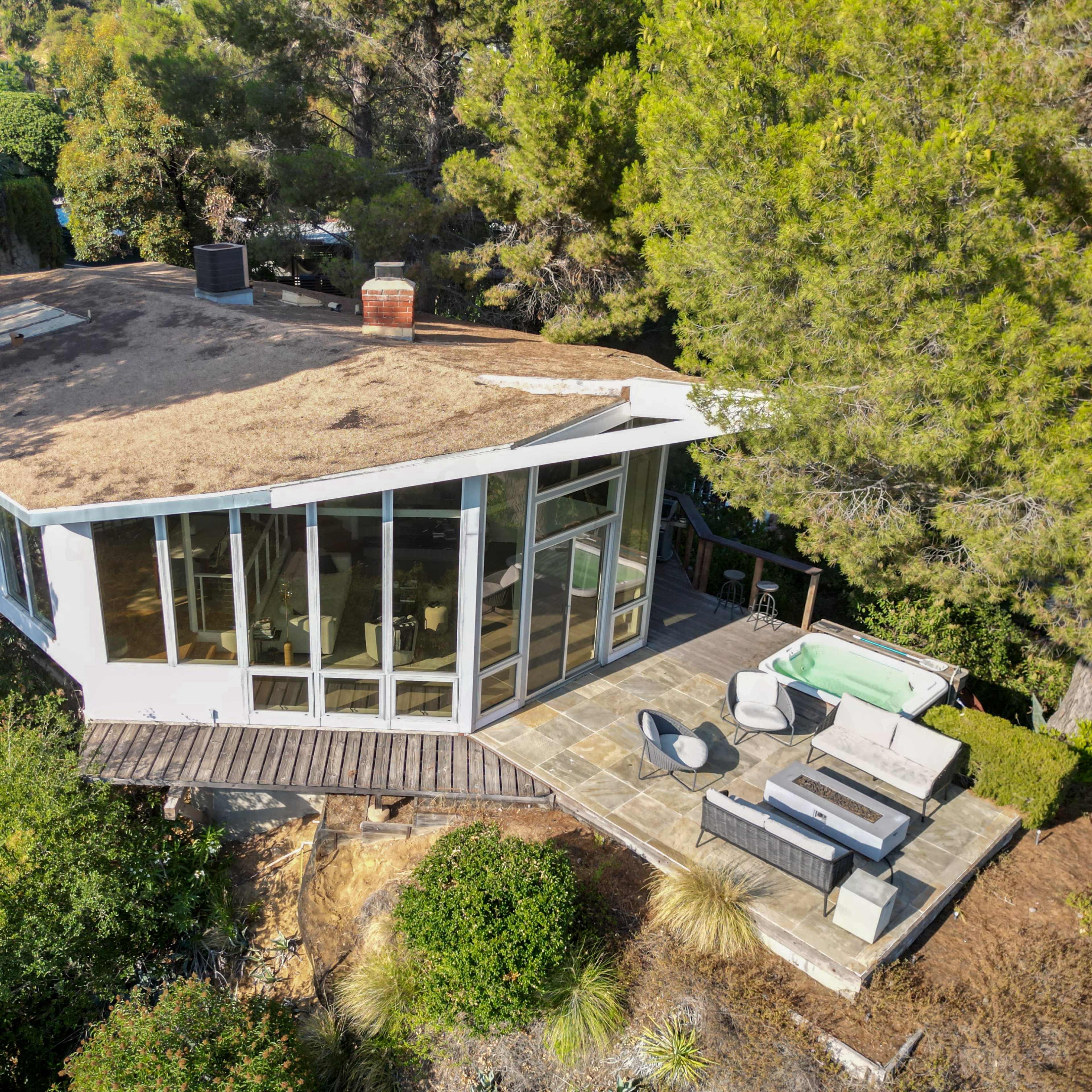 A modern flat-roofed house with large windows sits among trees, featuring a stone patio and outdoor seating area.
