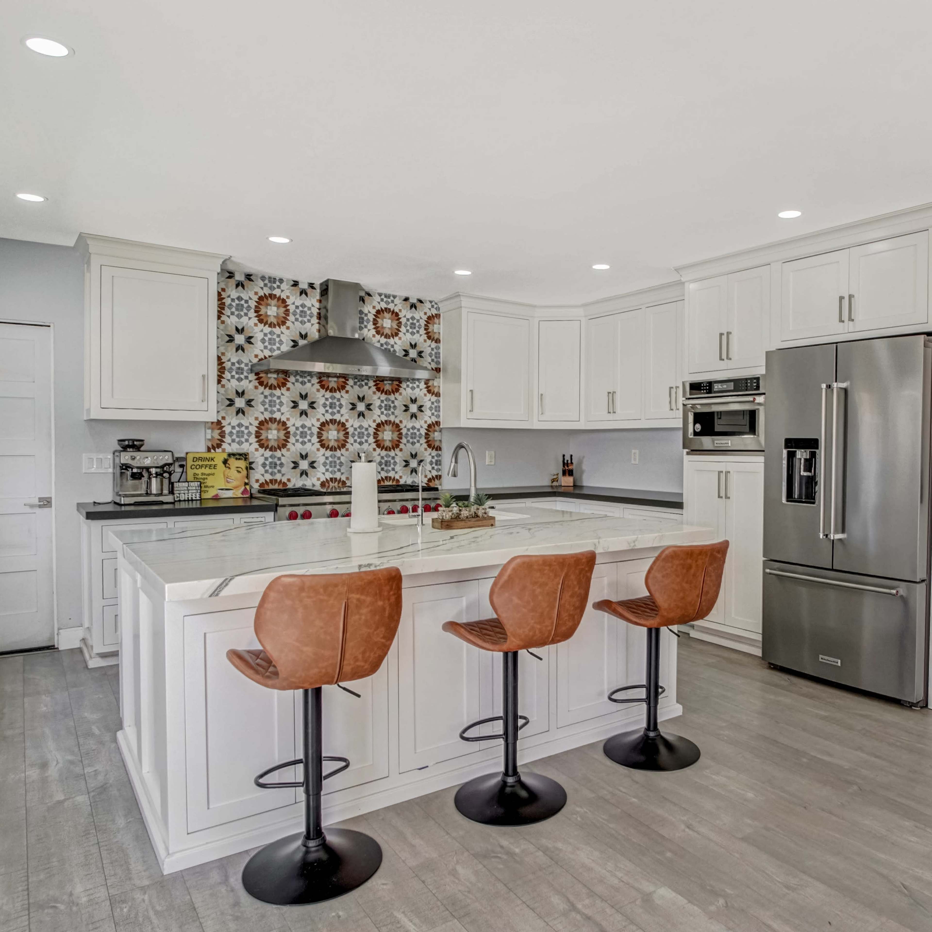 A modern kitchen features white cabinetry, a patterned tile backsplash, stainless steel appliances, and three brown bar stools at a large island.
