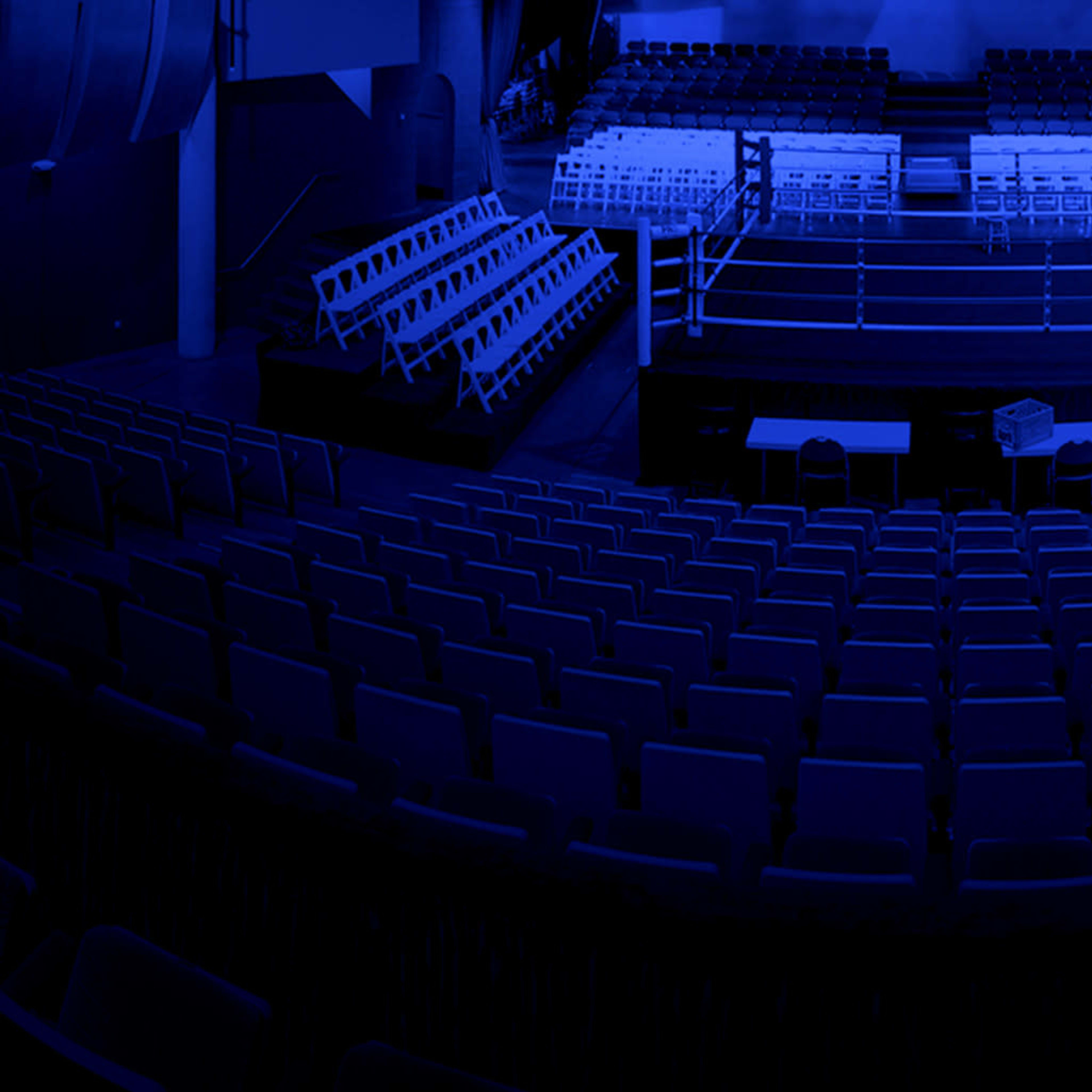 A boxing ring is set up in an empty arena, surrounded by rows of folding chairs under dim blue lighting.