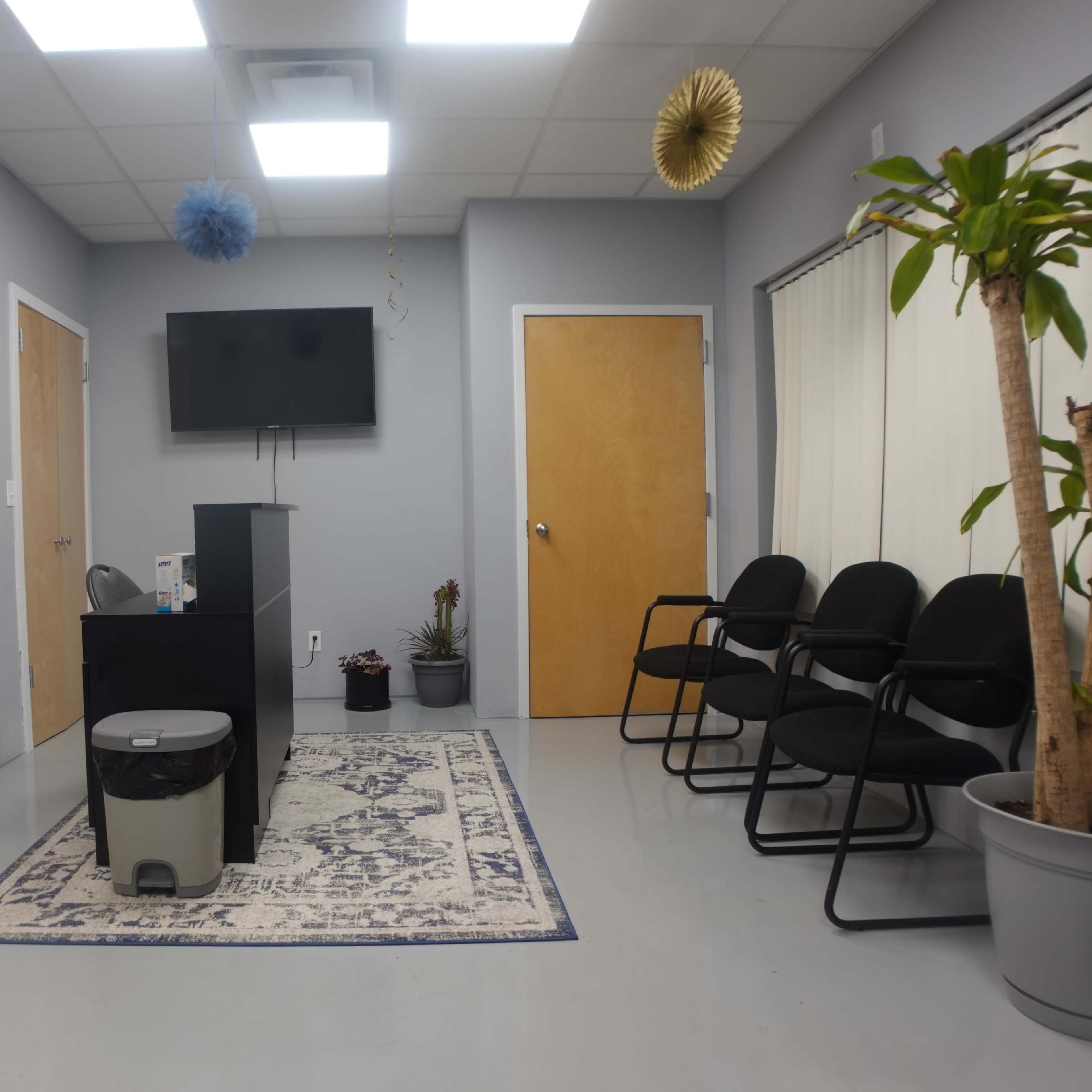 A waiting room with gray walls, a reception desk, chairs, a television, and a potted plant.