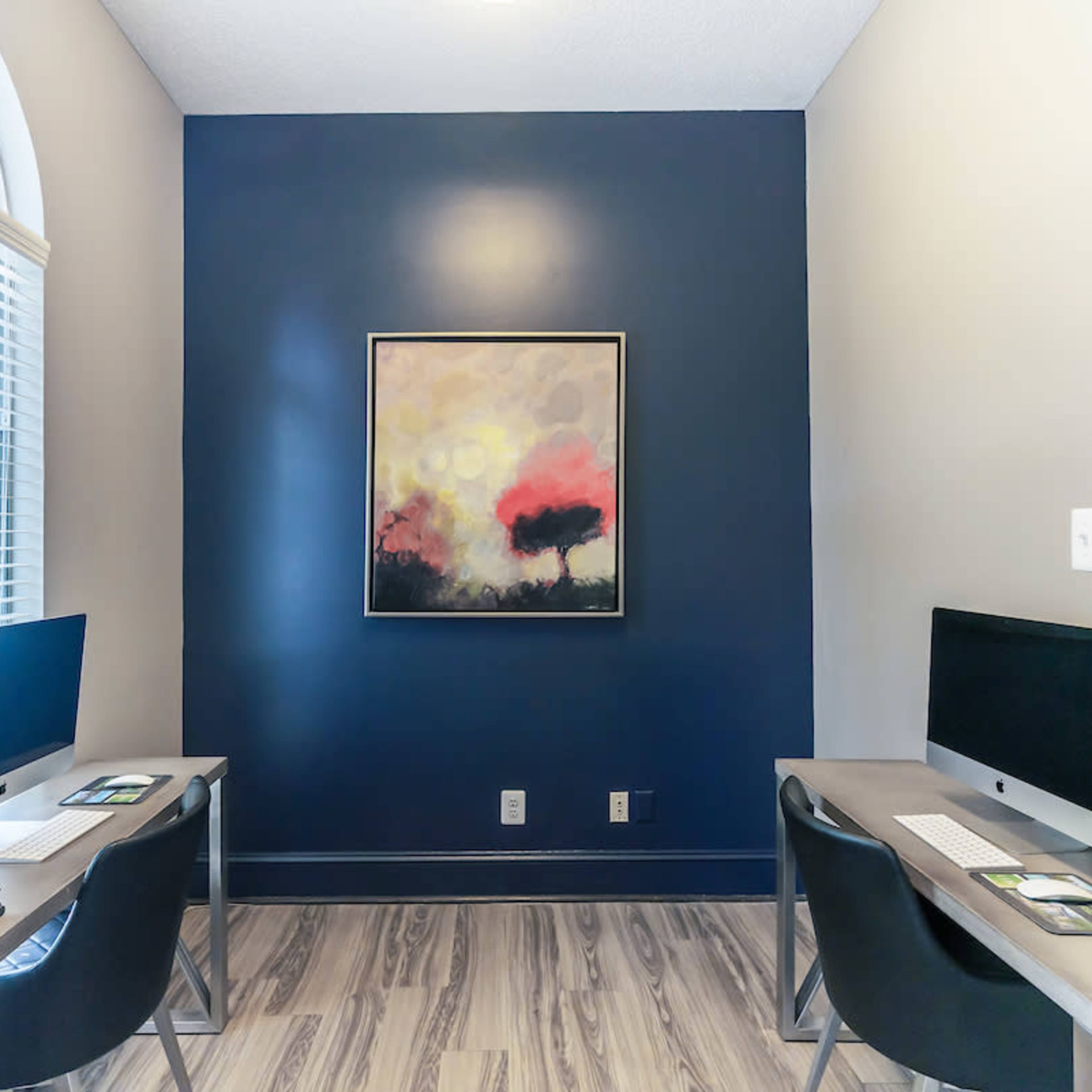 A workspace with two desks, each equipped with a computer, facing a dark blue wall that features a framed artwork above.