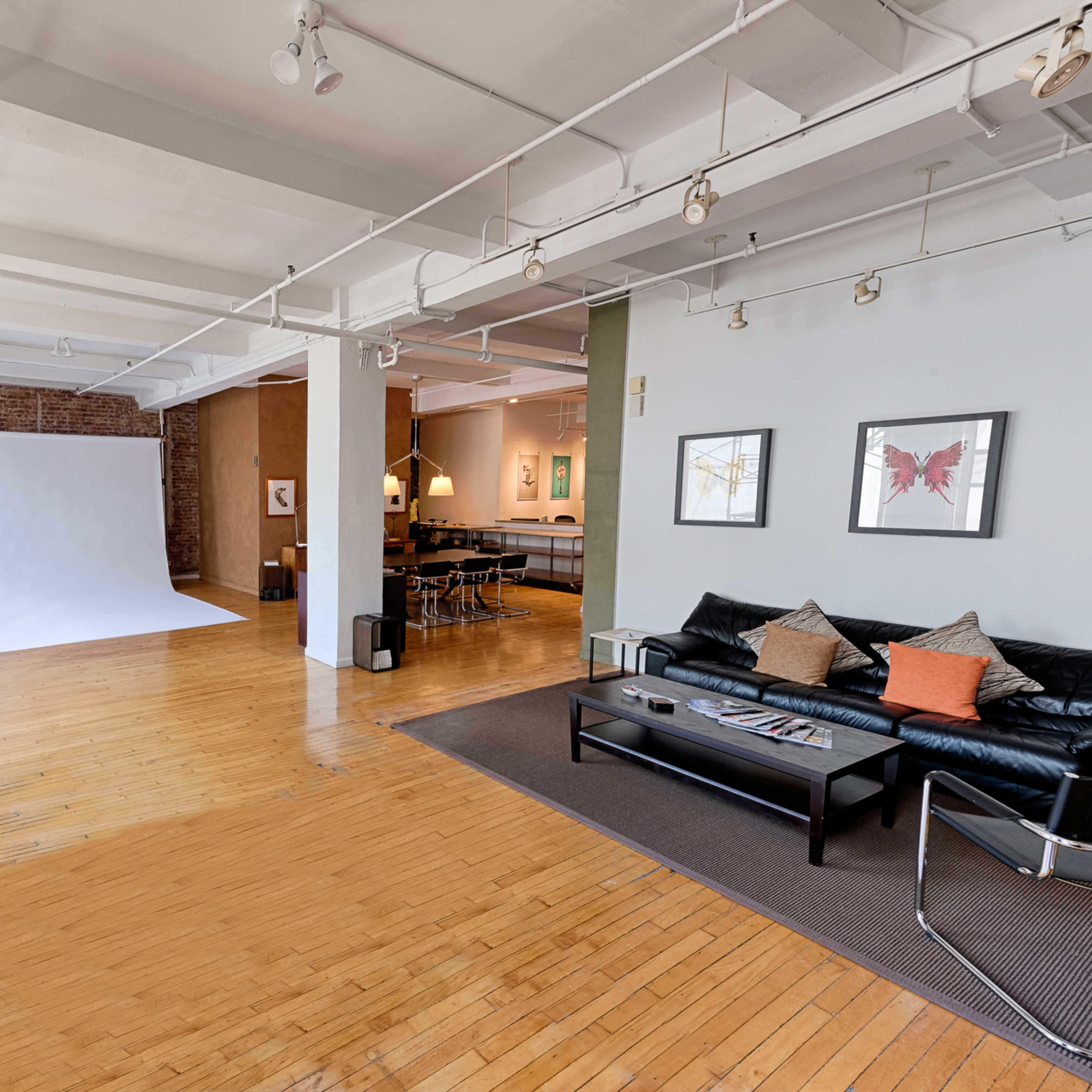 A spacious room with hardwood floors, featuring a white backdrop, a black leather couch, and modern furniture arranged for a cozy, open layout.