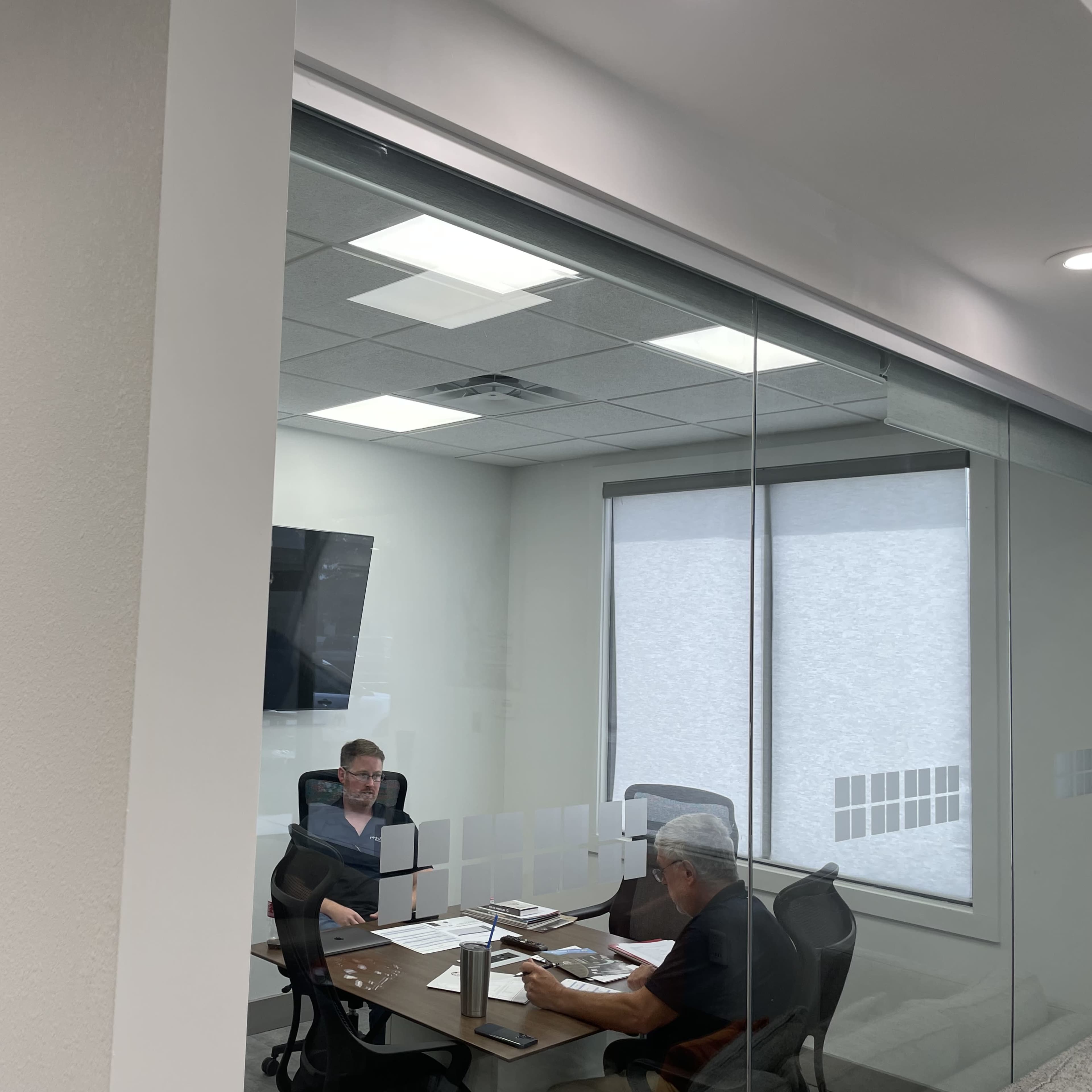 Two men are seated at a table in a conference room, discussing documents while a TV hangs on the wall.