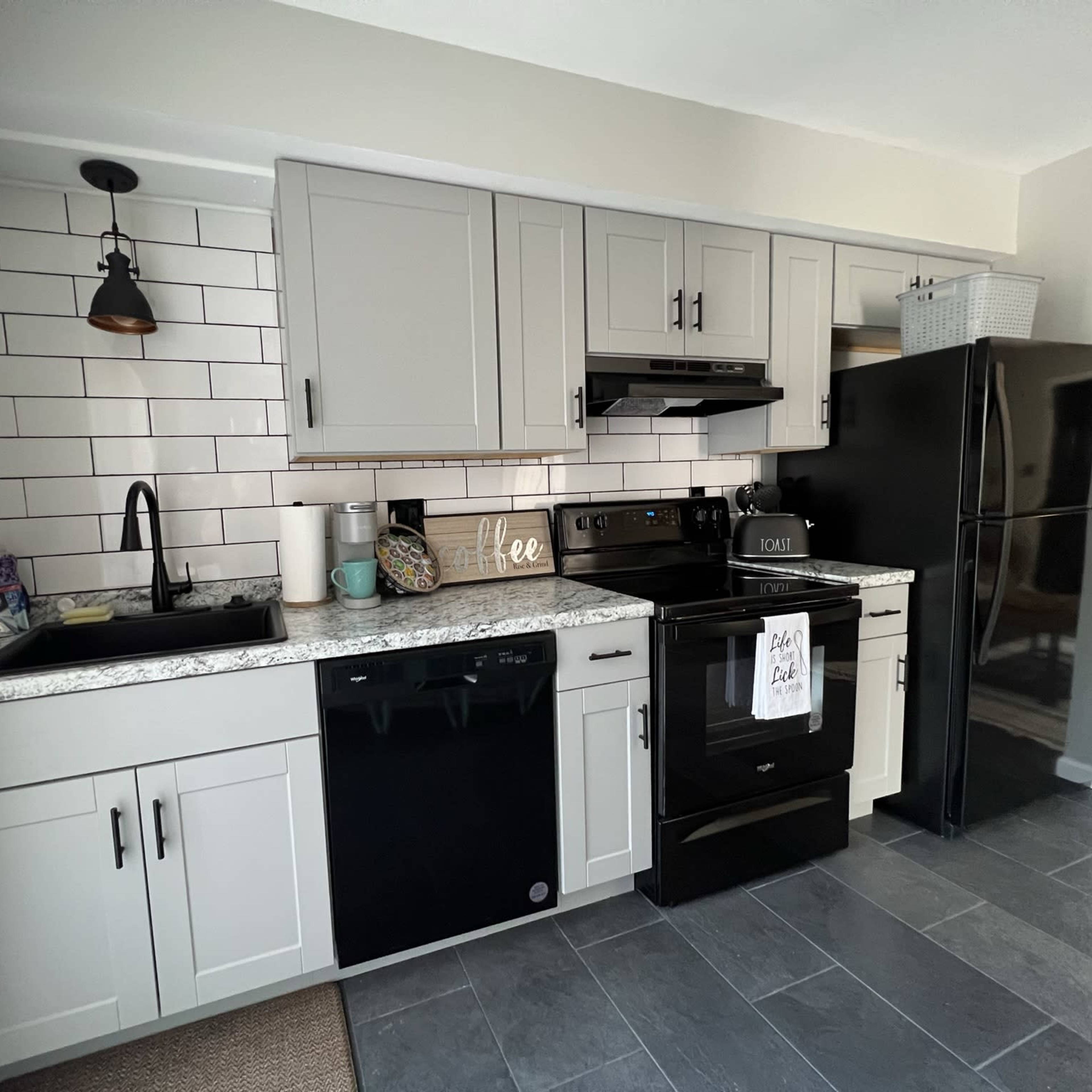 A modern kitchen with gray cabinetry, a black refrigerator, and dark appliances, featuring a white tile backsplash and granite countertops.