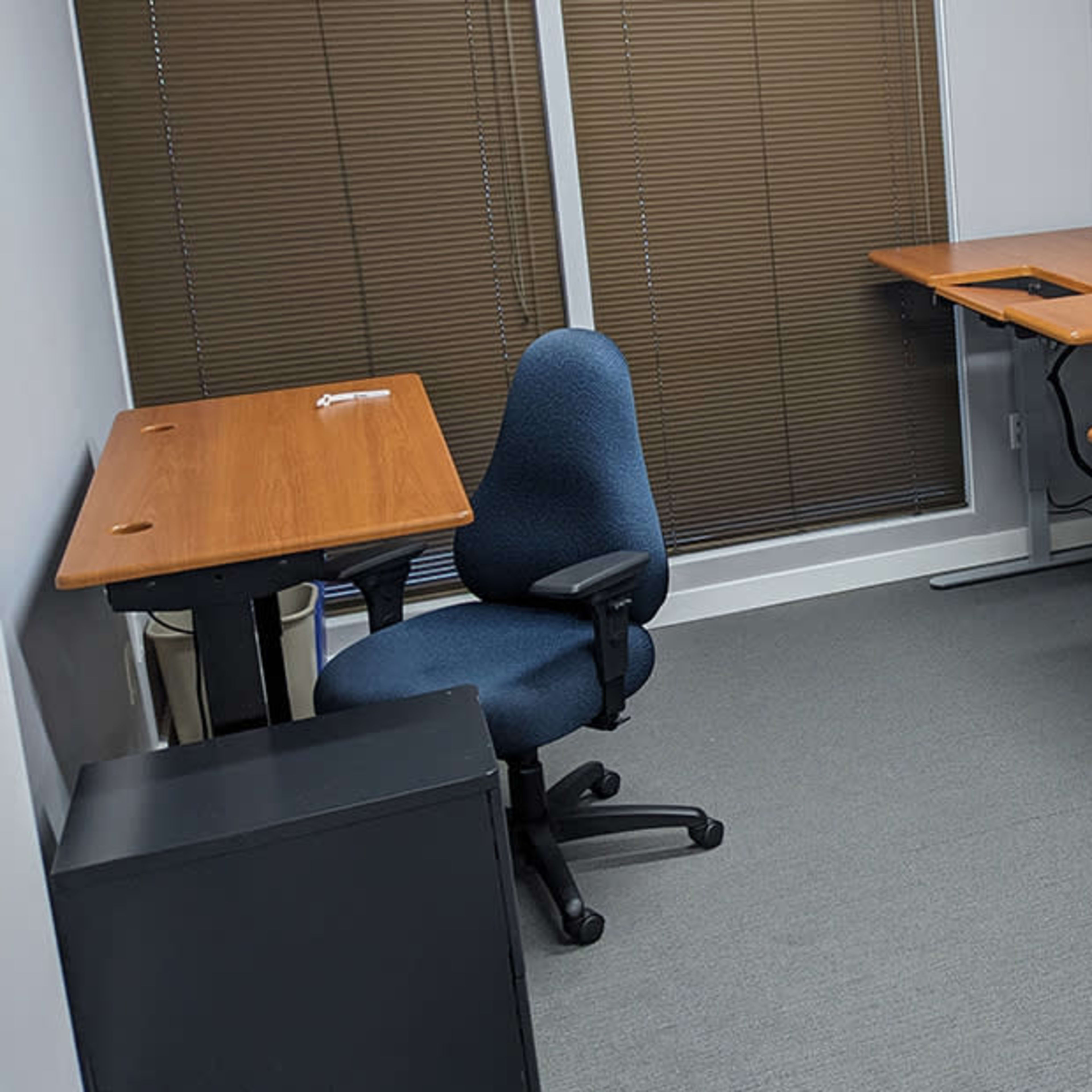A small office space with three wooden desks and a blue office chair arranged in a well-lit area with blinds on the windows.