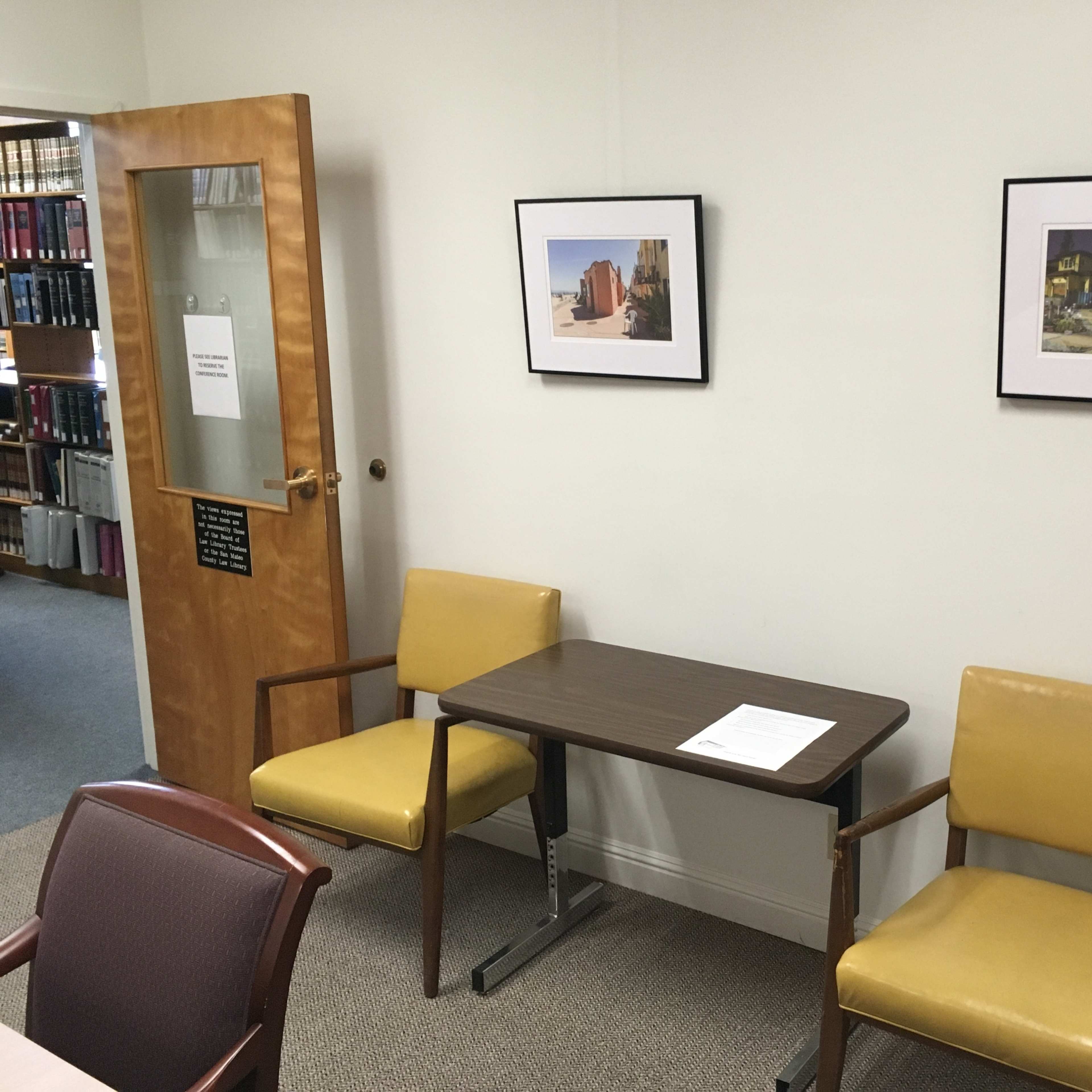 A small room with two yellow chairs, a wooden table, and two framed pictures hanging on the wall.