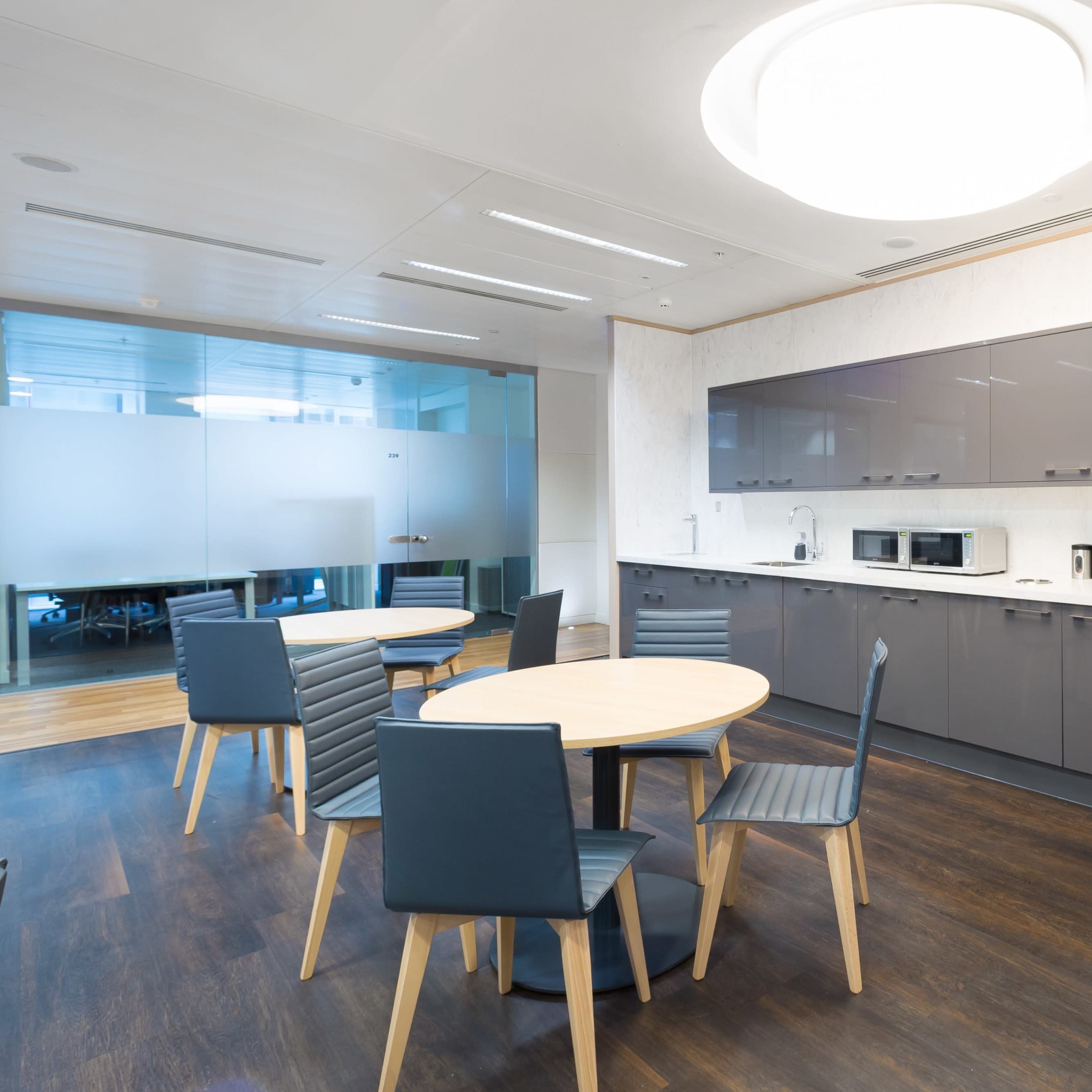 A modern office kitchen area features a circular table with six chairs, gray cabinetry, a coffee machine, and two microwaves against a light-colored wall.