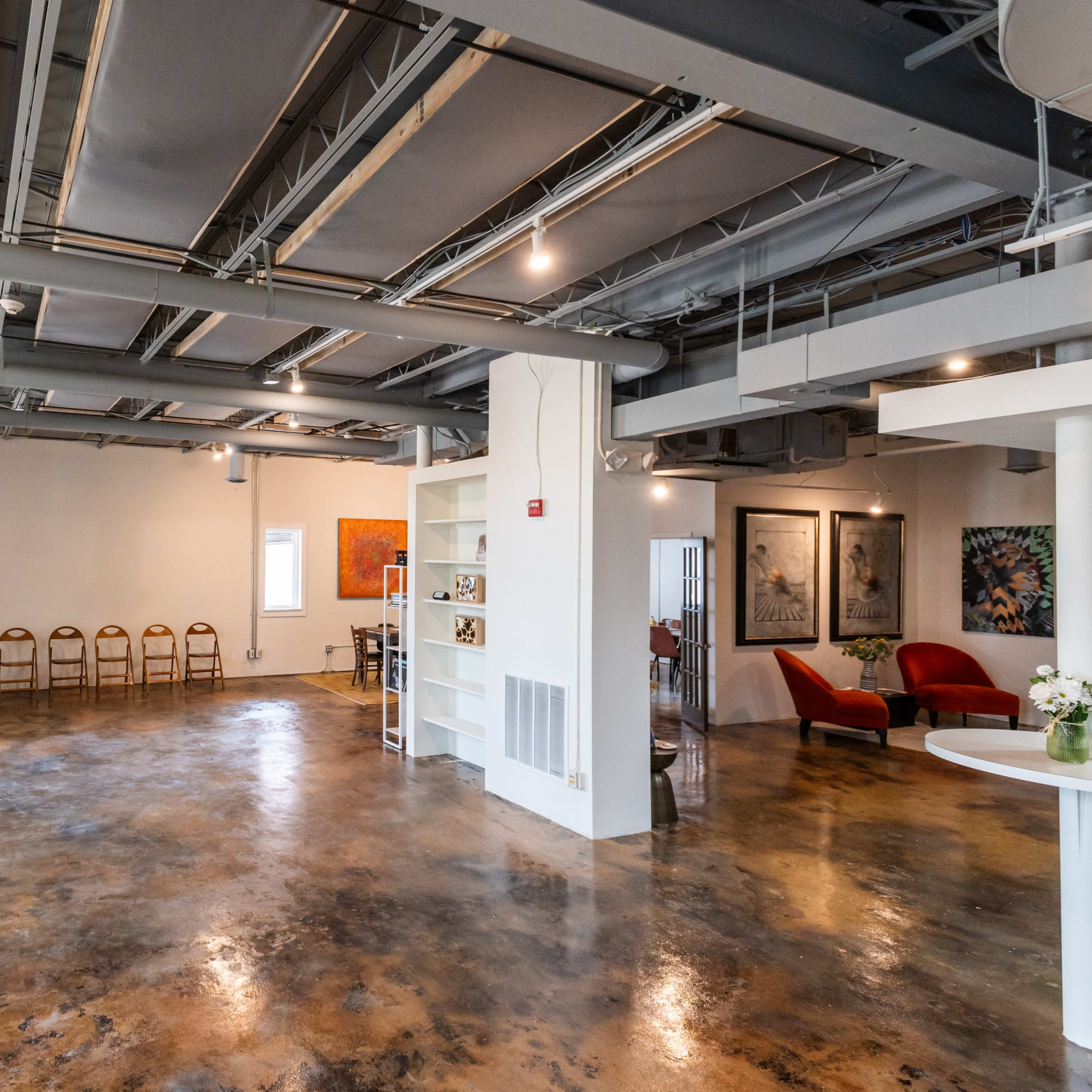 A spacious indoor area with a polished concrete floor, featuring several chairs arranged along the walls and artwork hanging on the white walls.