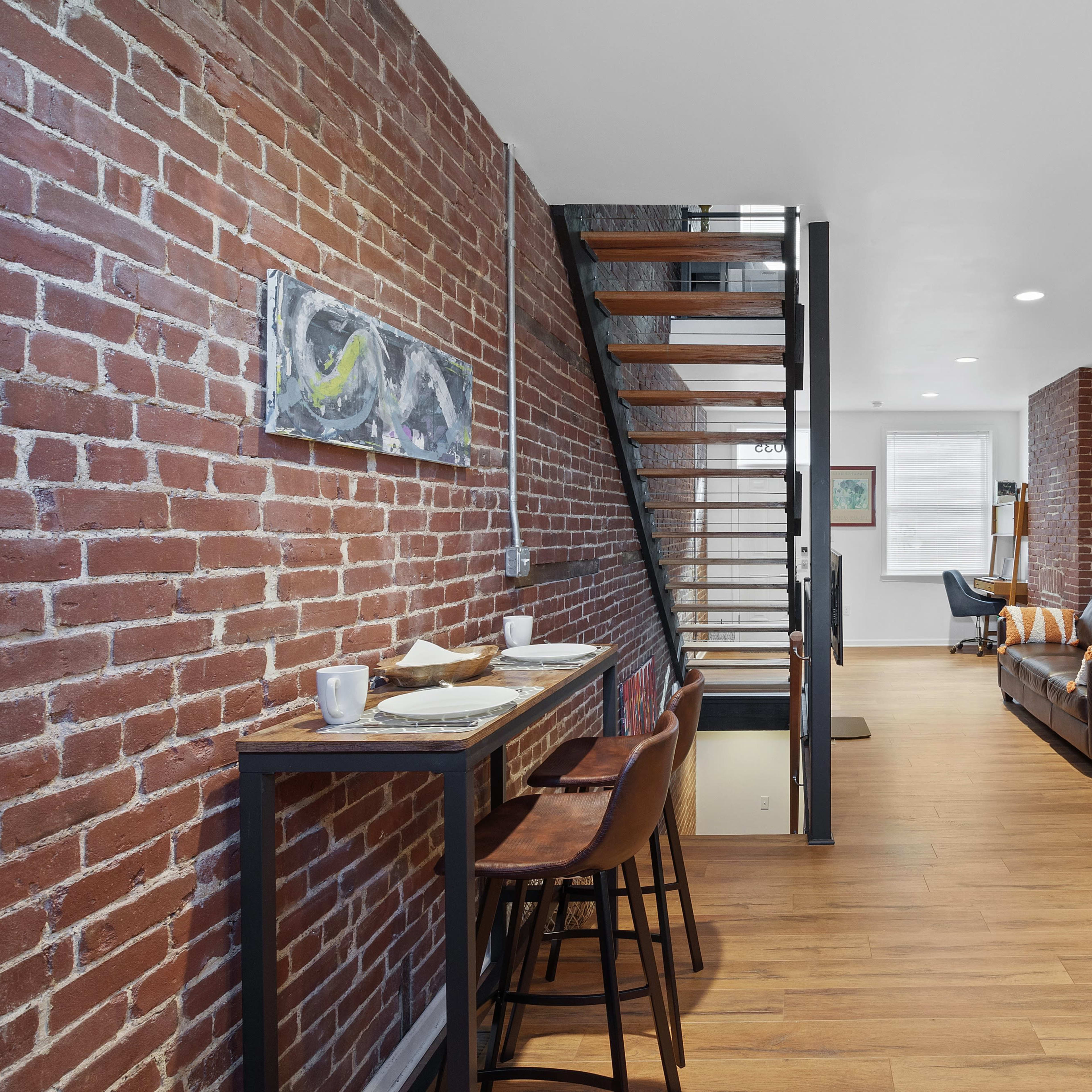 A modern kitchen and living area featuring exposed brick walls, a staircase, and contemporary furniture.