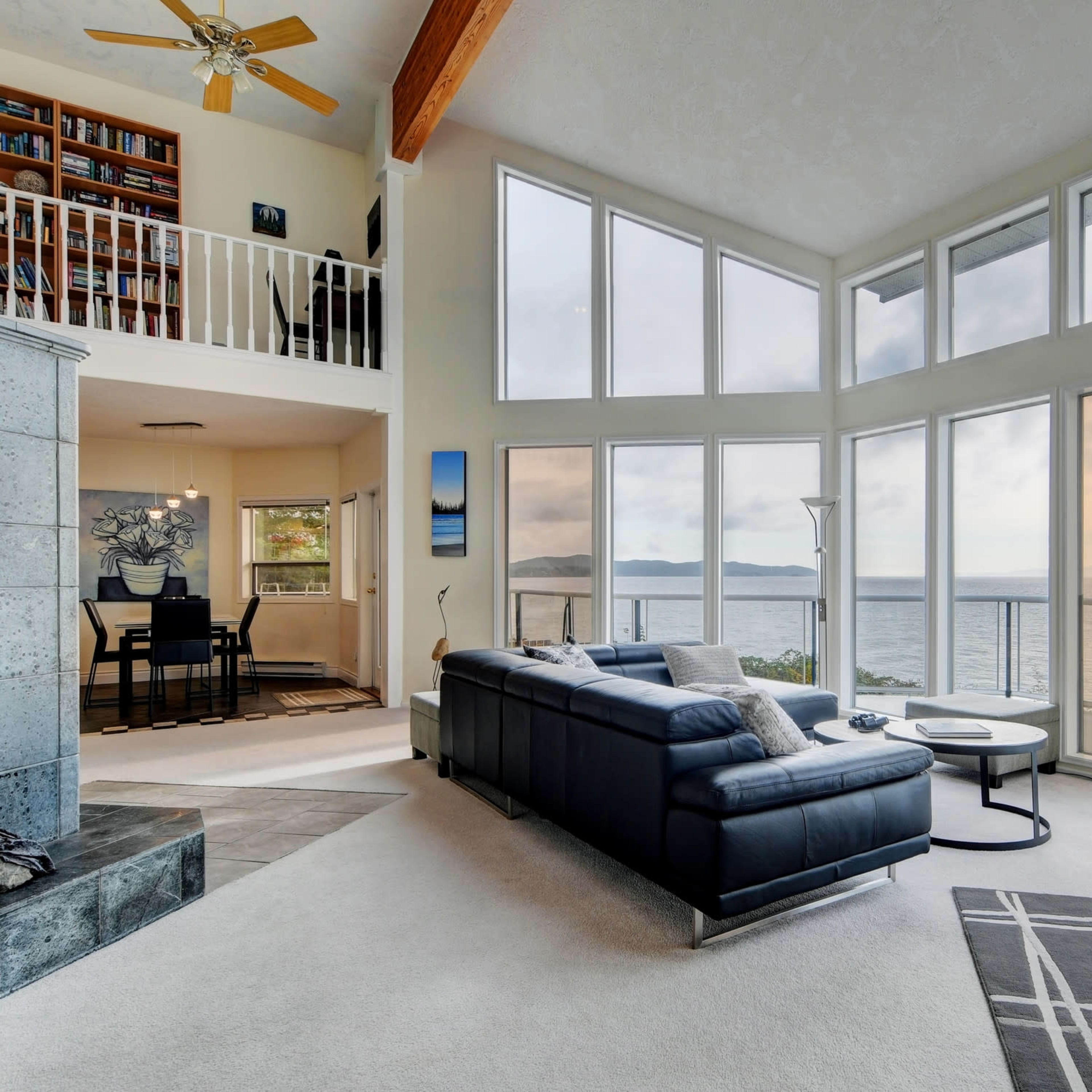 A modern living room features large windows overlooking the ocean, with a sectional sofa, coffee table, and a bookshelf in the background.