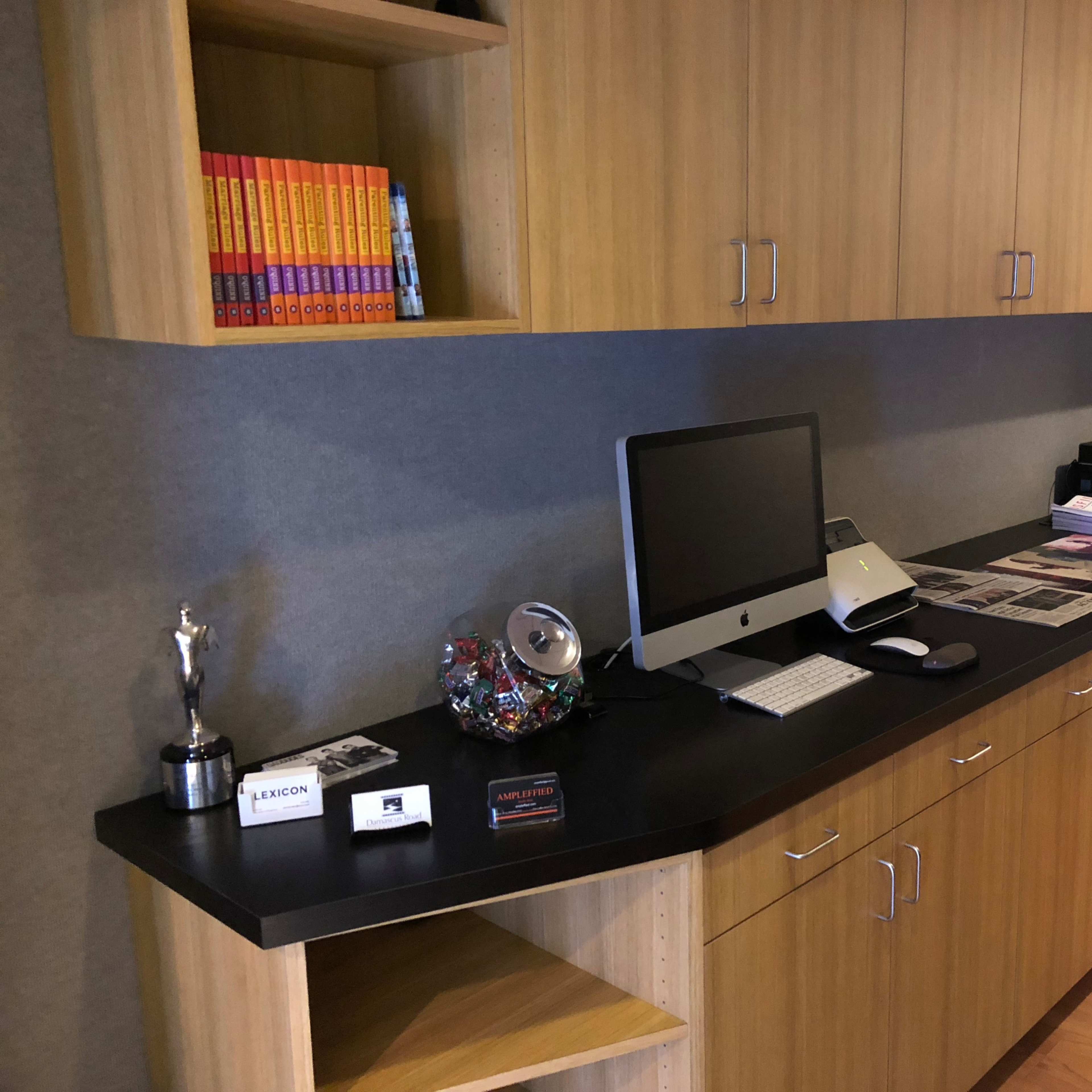 A wooden office space featuring a black countertop with a computer, printer, and various books and awards displayed on shelves.