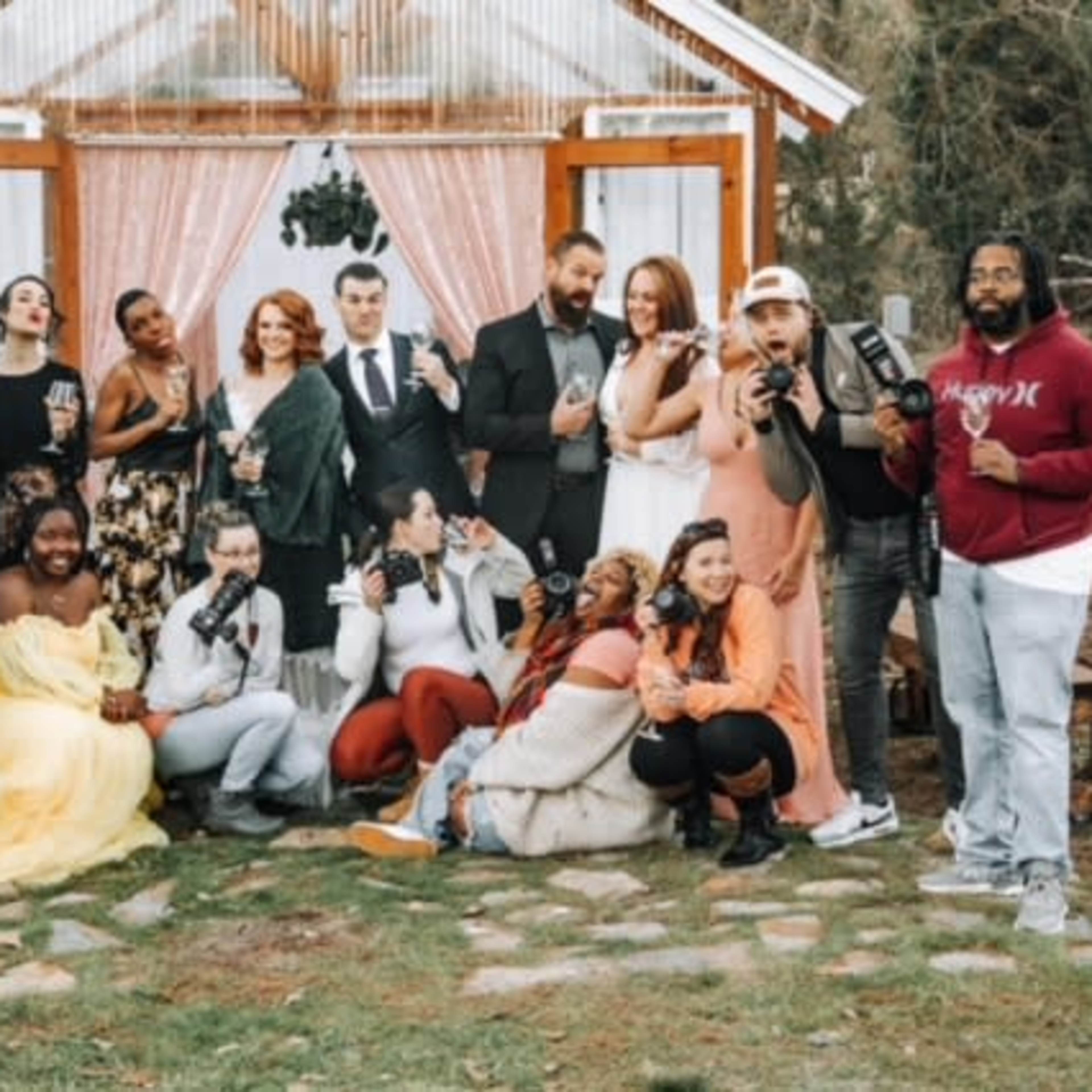 A group of people poses together in front of a decorated outdoor backdrop, with some holding drinks and cameras.