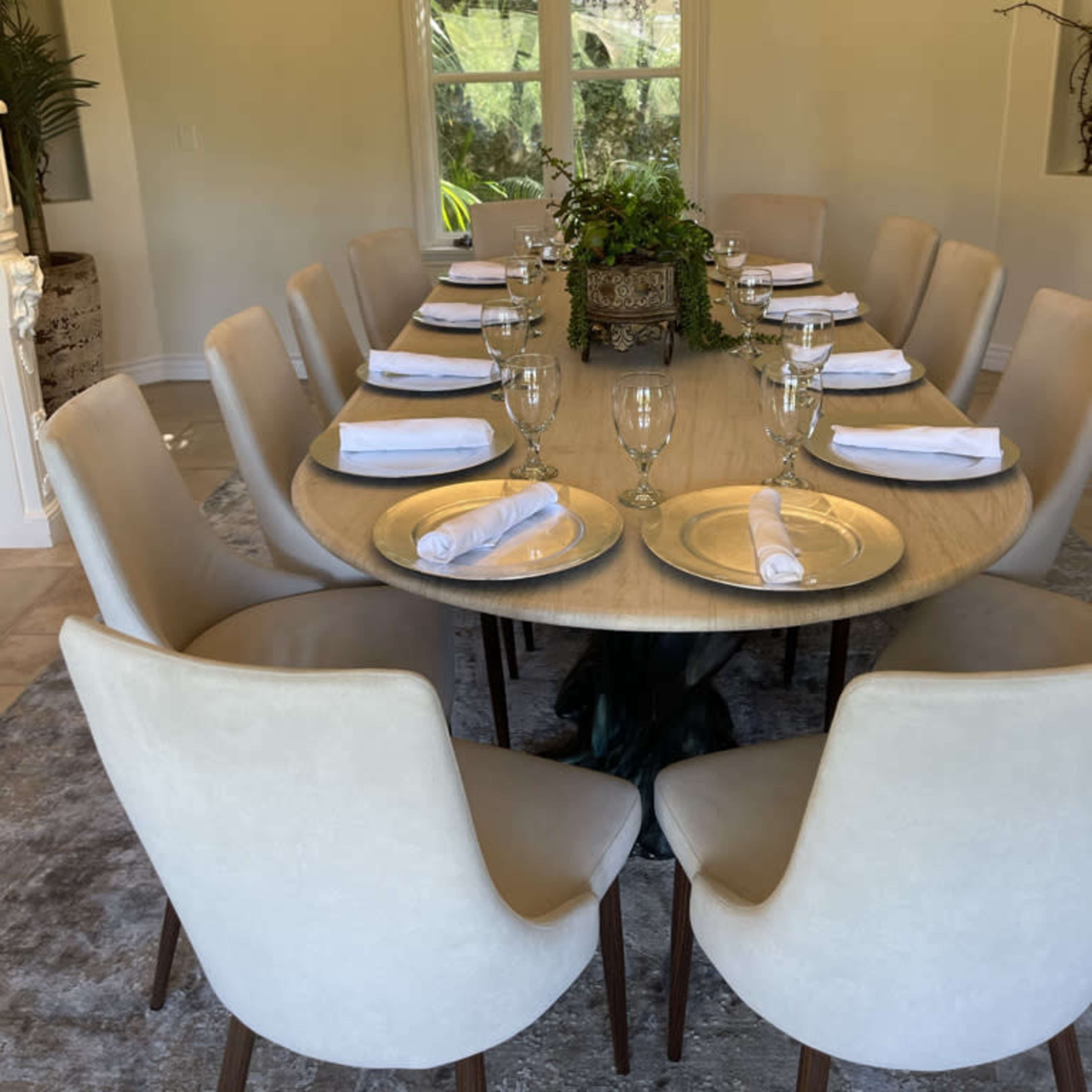 A round dining table set with plates, napkins, and glasses is arranged in a well-lit room with large windows and greenery visible outside.
